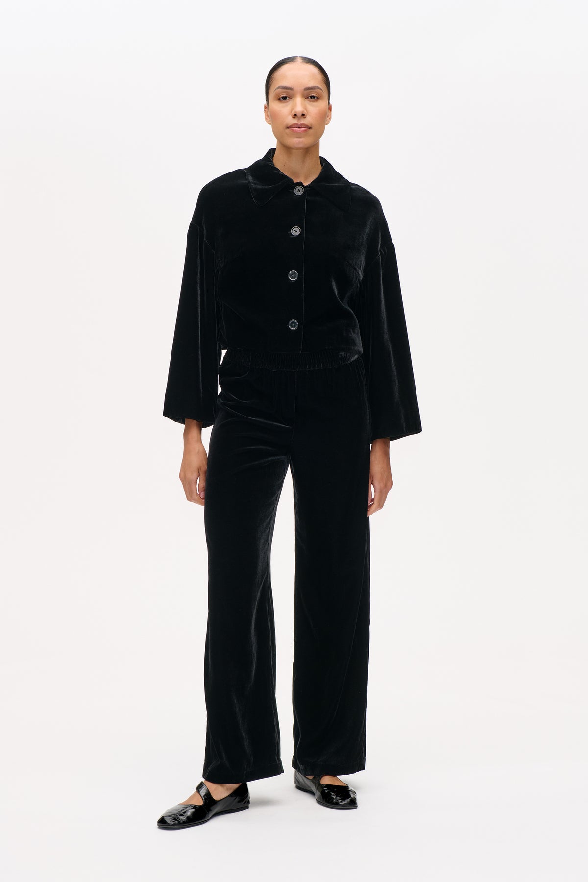 Front view of a model wearing a black velvet button-front jacket with a wide collar and matching wide-leg trousers.