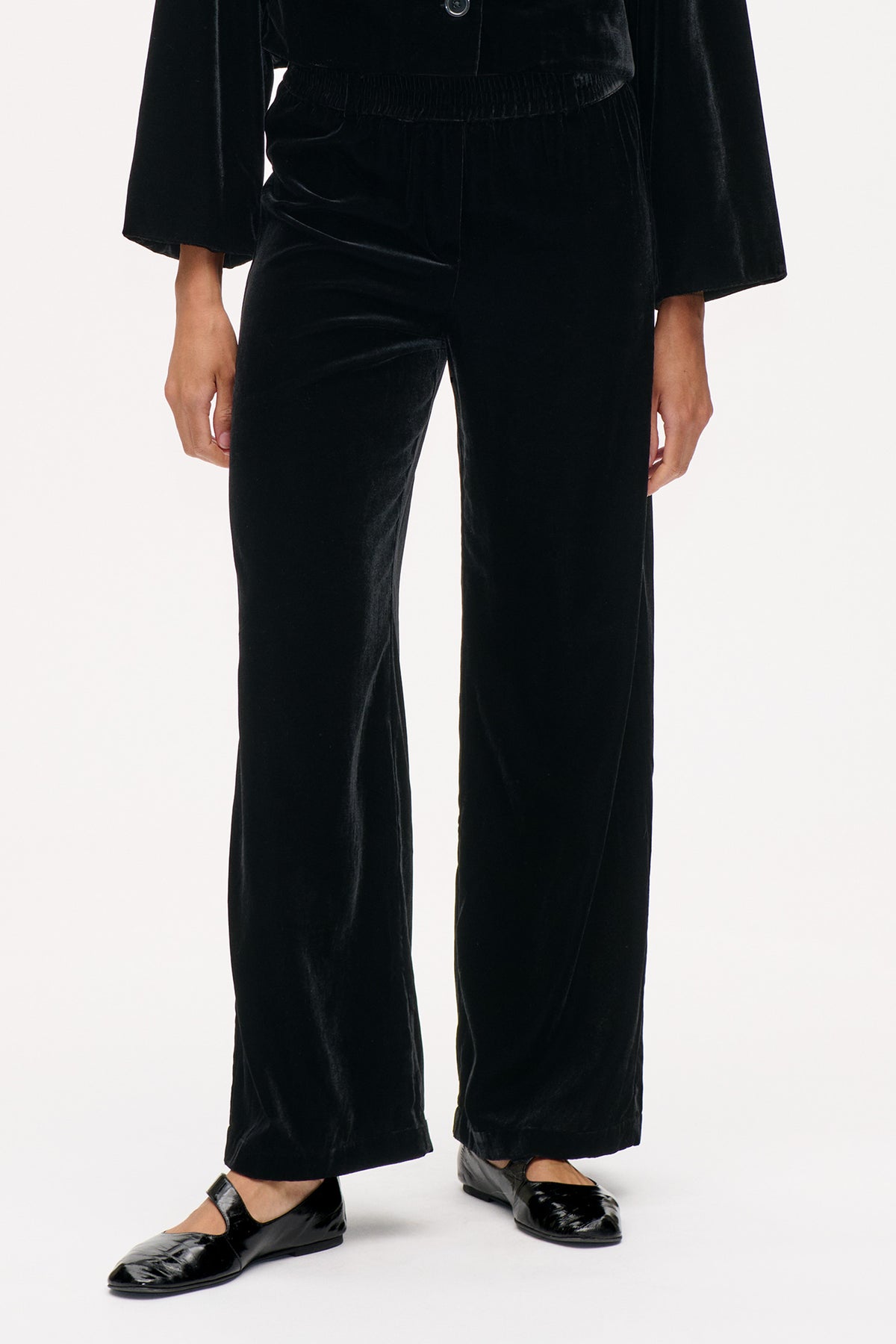 Front detail of black velvet wide-leg trousers with a gathered waistband, styled with a black jacket and glossy Mary Jane flats.