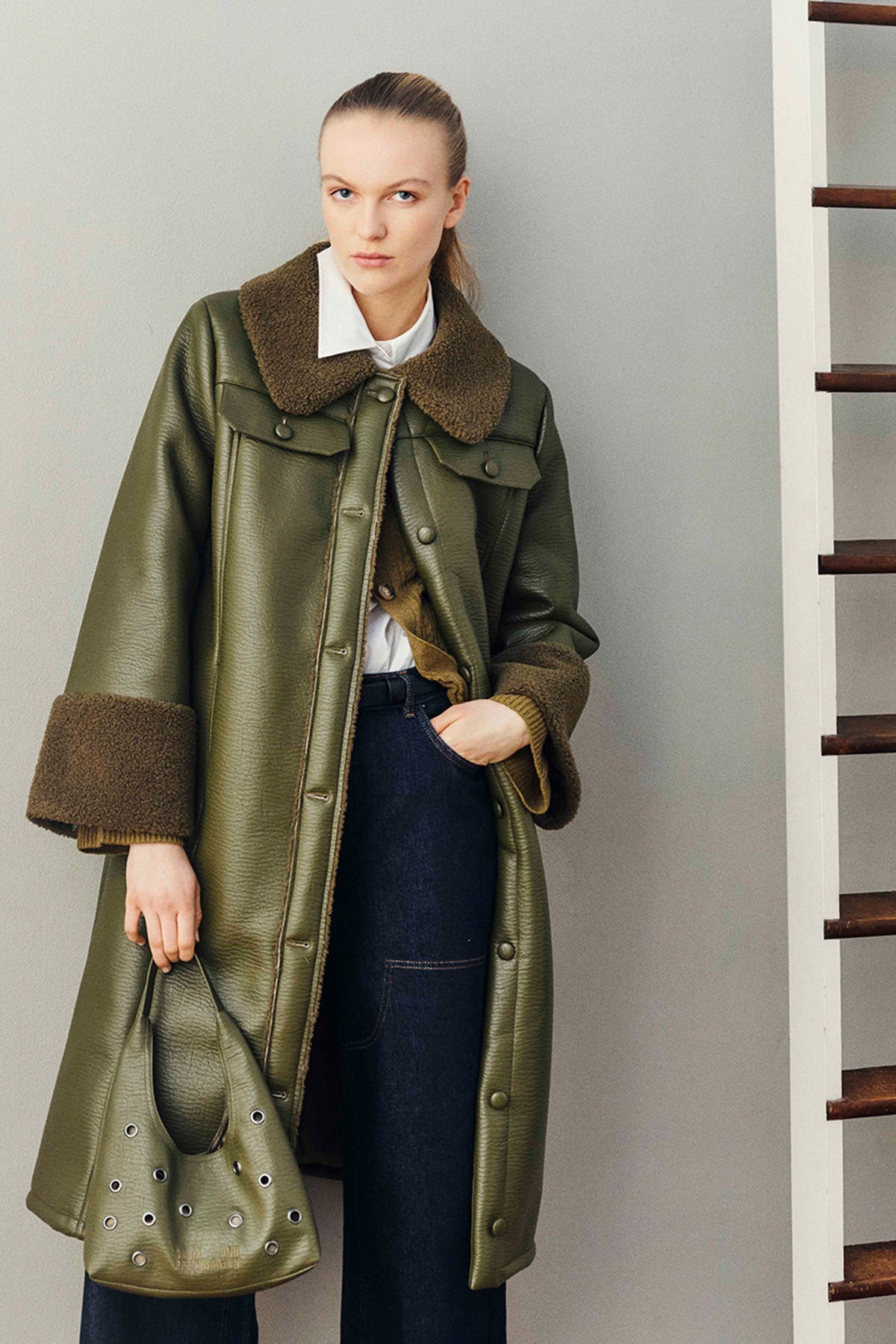 Model in an olive green textured jacket with a brown plush collar and cuffs, white shirt and dark jeans, holding a matching eyelet bag.