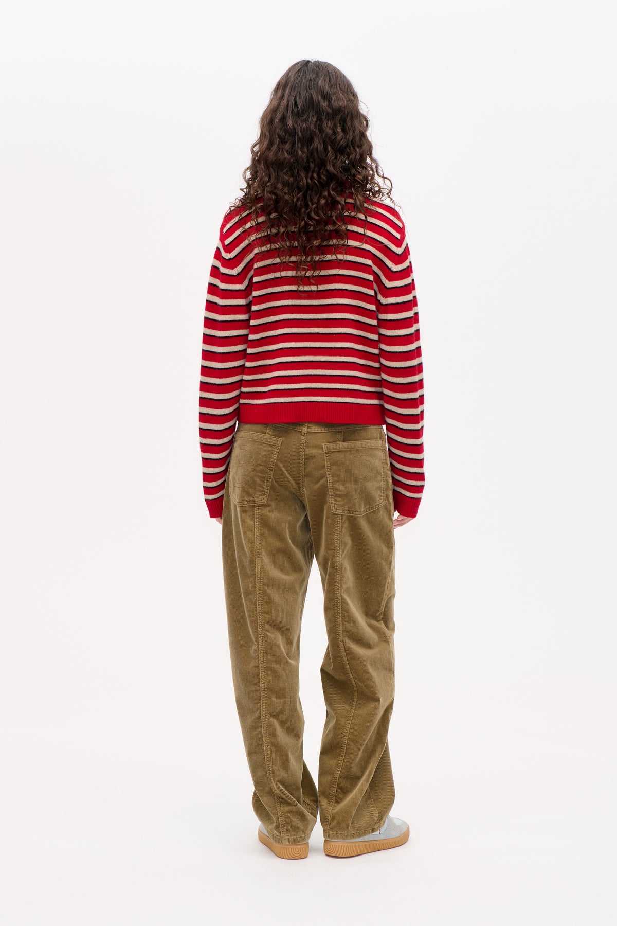 Achteraanzicht van een model met een rode trui met crème- en marineblauwe horizontale strepen en een losse bruine ribfluwelen broek.