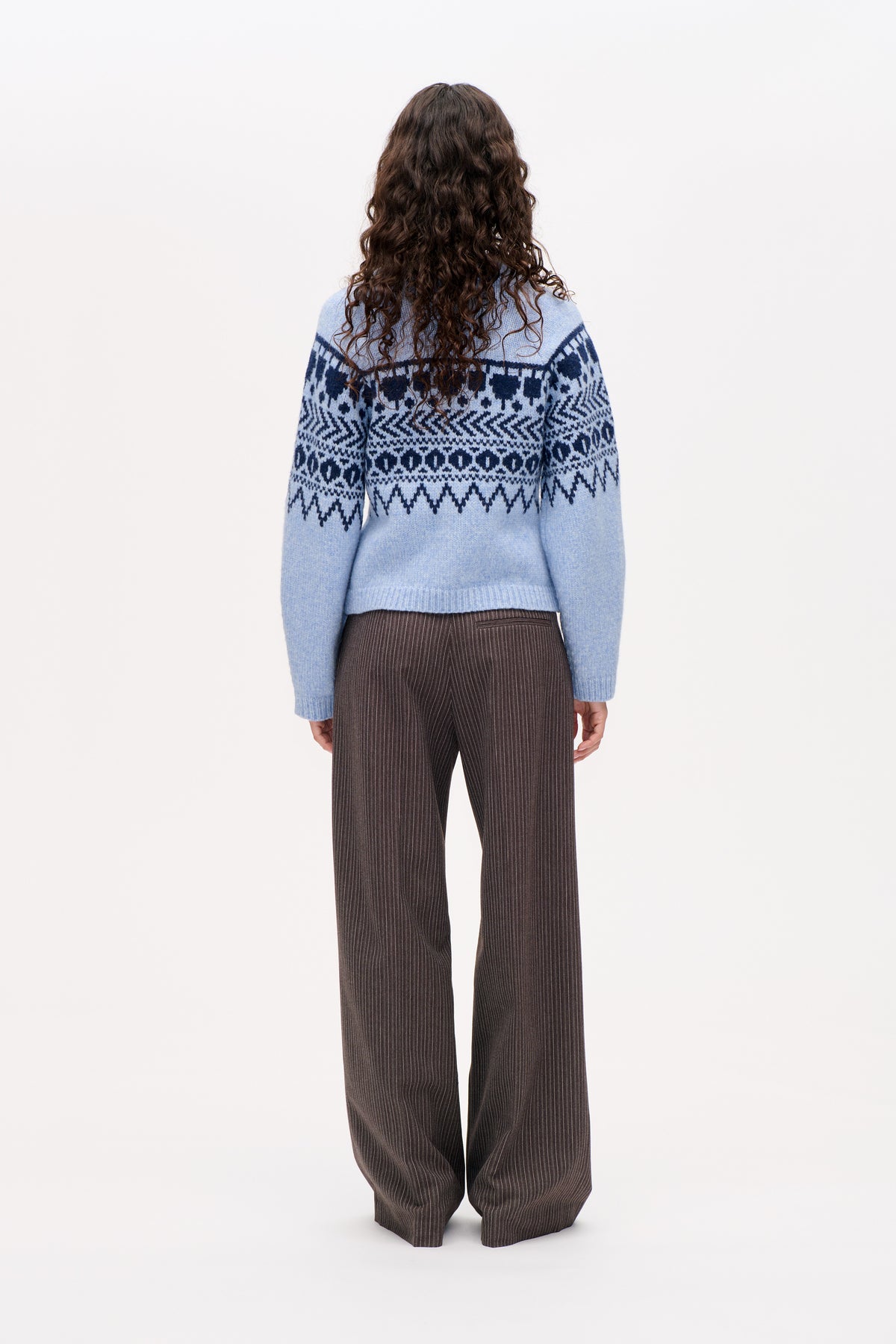 Back view of a light blue sweater with dark navy geometric Fair Isle patterns, paired with dark brown pinstriped wide-leg trousers.