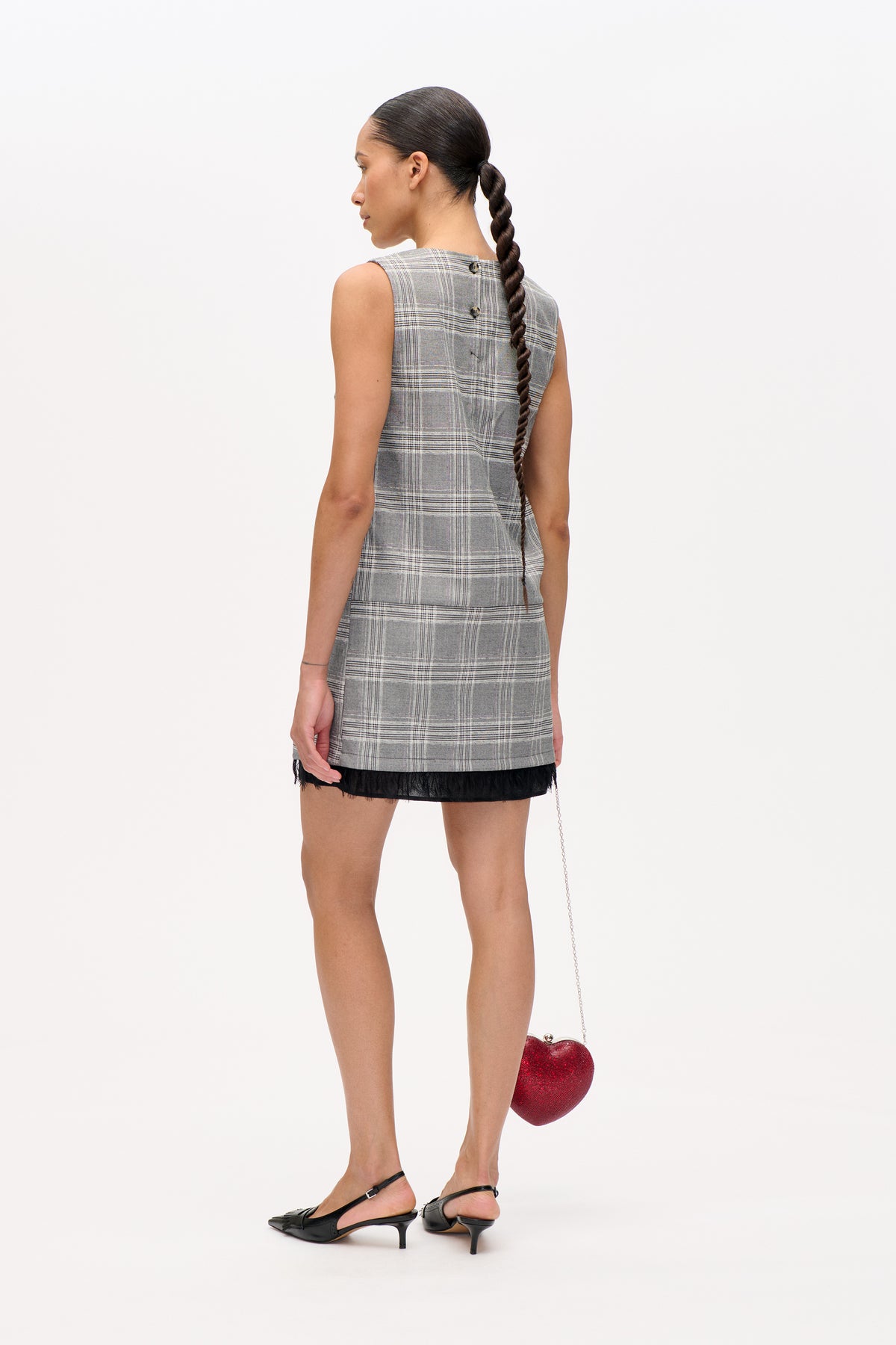 Back view of a model wearing a sleeveless grey plaid mini dress with two buttons and a black sheer hem.