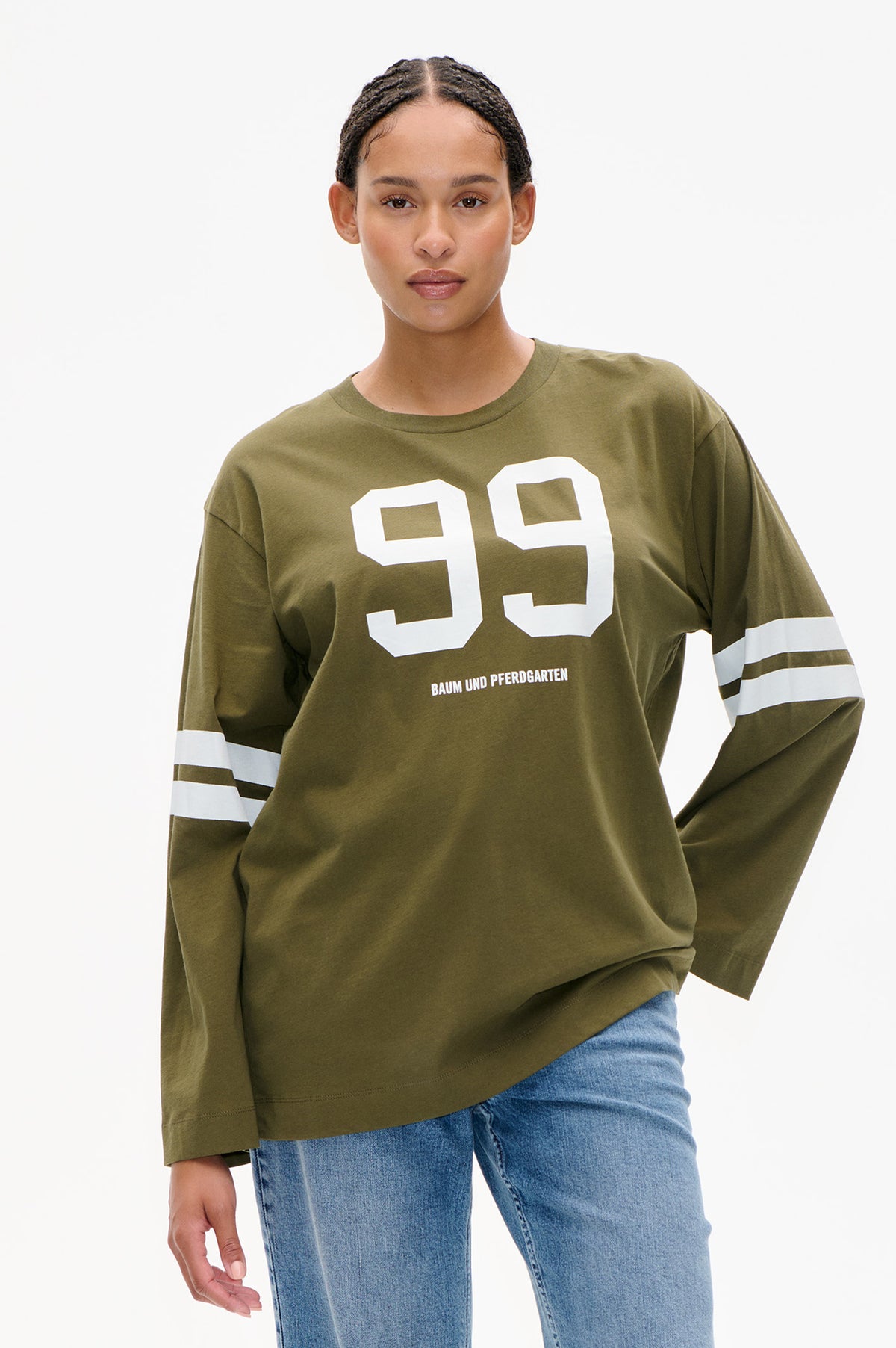 Front view of an olive-green long-sleeved T-shirt with a large white 99 graphic, Baum und Pferdgarten lettering and white sleeve stripes, paired with blue jeans.