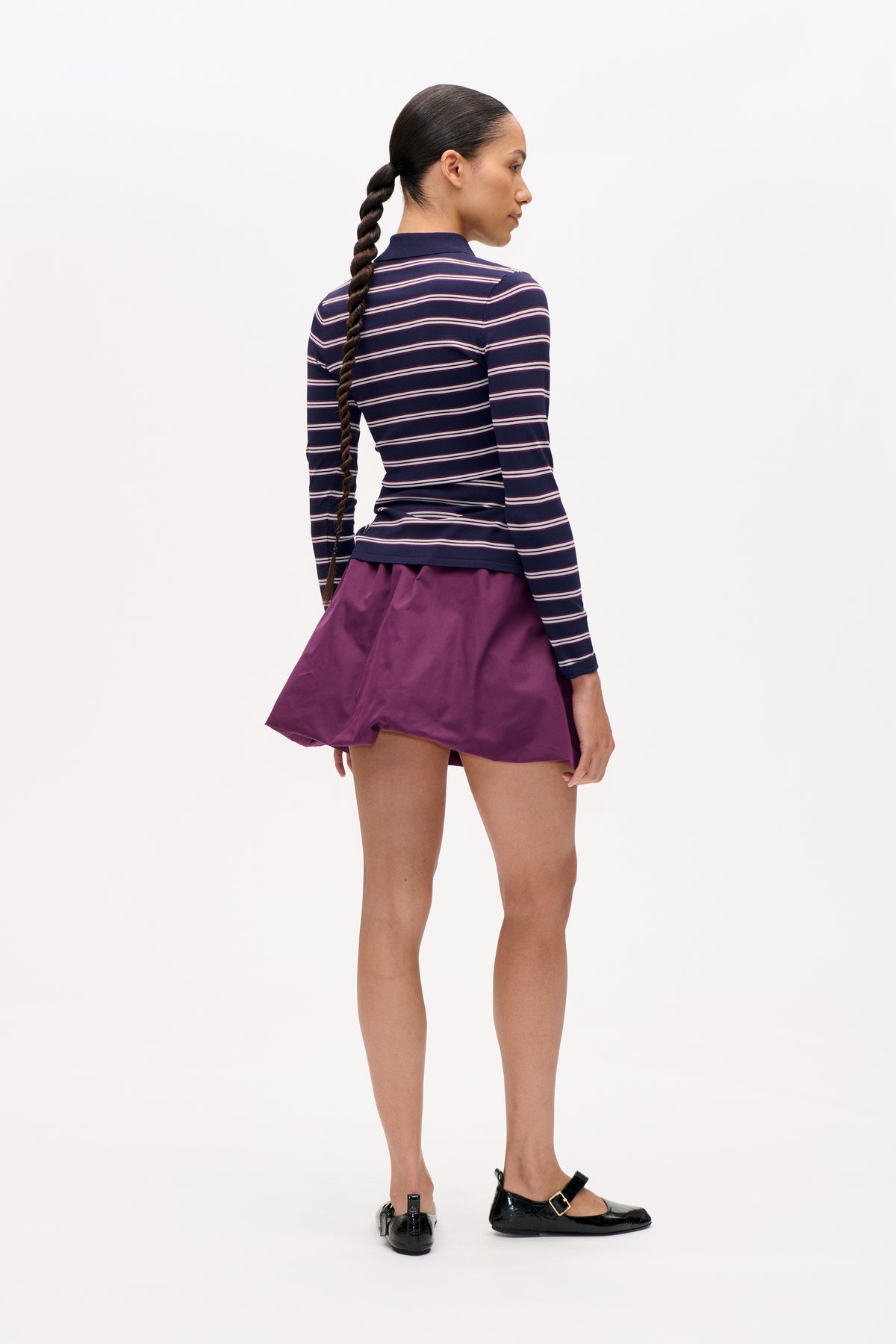 Model in a rear three-quarter view wearing a navy long-sleeved striped collared top, plum skirt and black buckle flats.