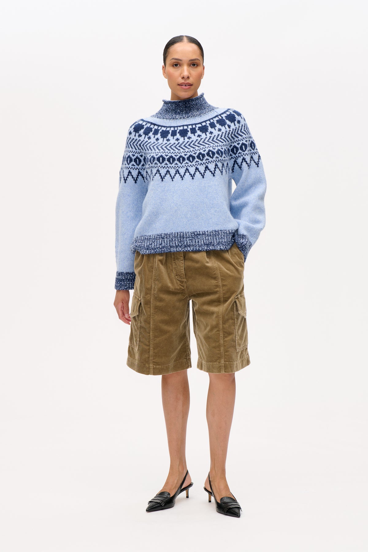 Vooraanzicht van een model in een lichtblauwe coltrui met marineblauwe geometrische banen en zandkleurige ribfluwelen shorts.