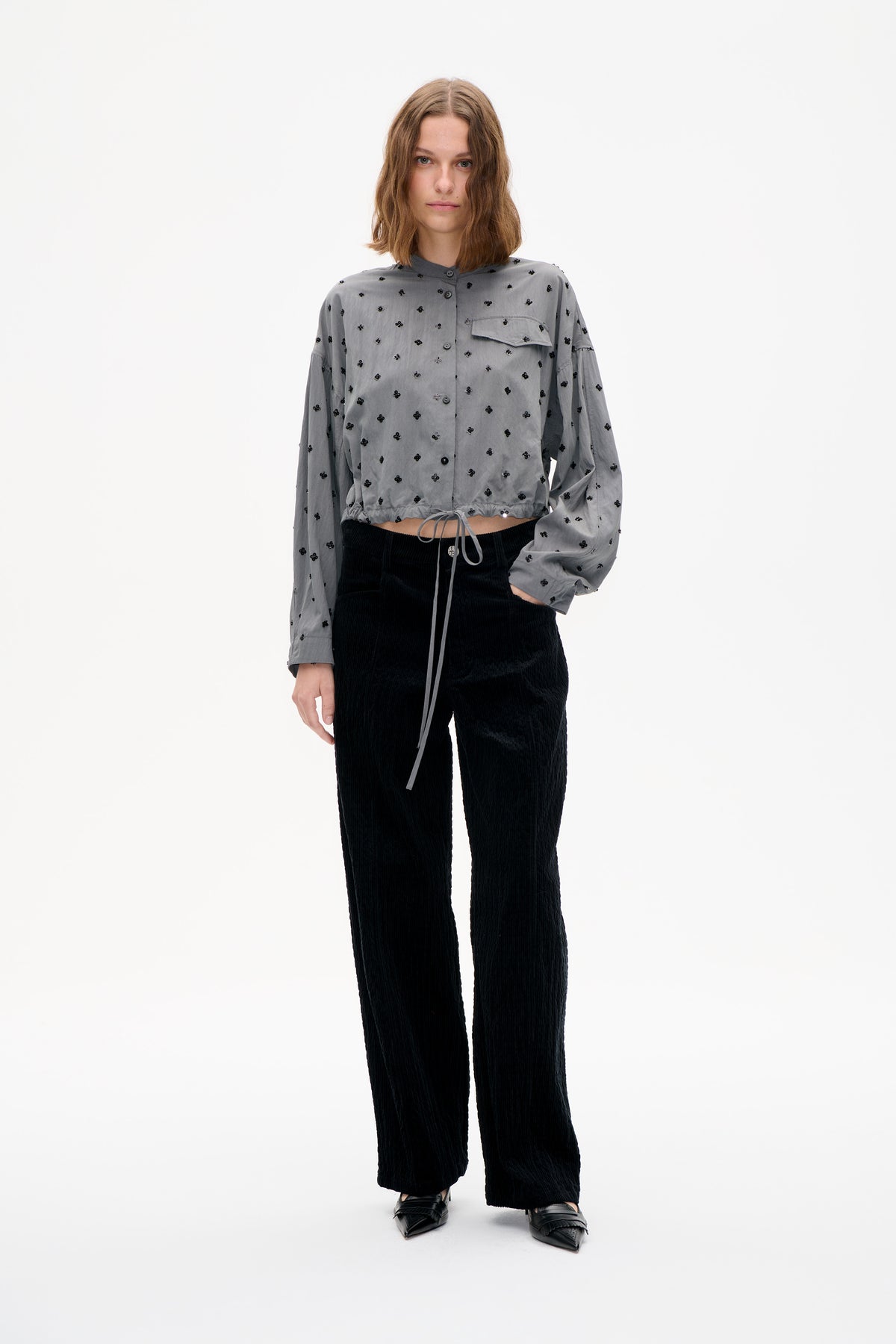 Front view of a model wearing a cropped grey button-up shirt with black floral embellishments, chest pocket and drawstring hem, paired with black trousers.
