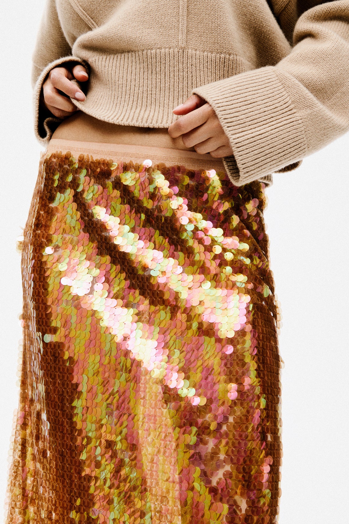 Close-up campaign crop of a person lifting a beige rib-knit sweater above a skirt covered in multicoloured sequins.