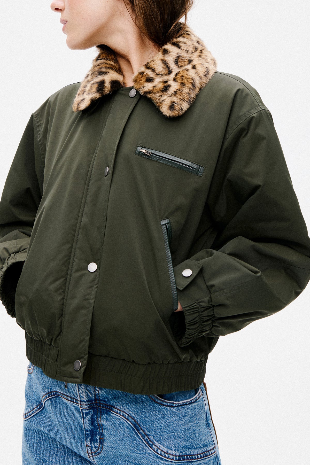 Close-up campaign view of a model wearing a dark olive jacket with a tan-and-black animal-print collar, zip chest pocket and snap buttons, paired with blue jeans.