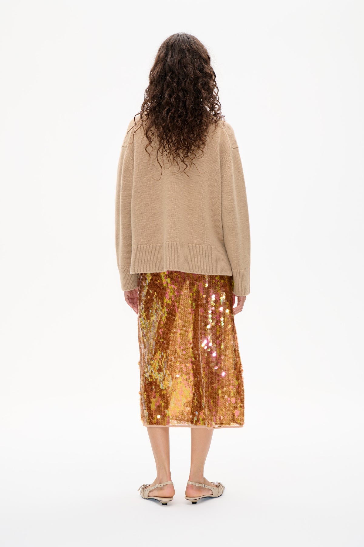 Back view of a model wearing a beige knit sweater and a knee-length skirt covered in shimmering multicoloured sequins.