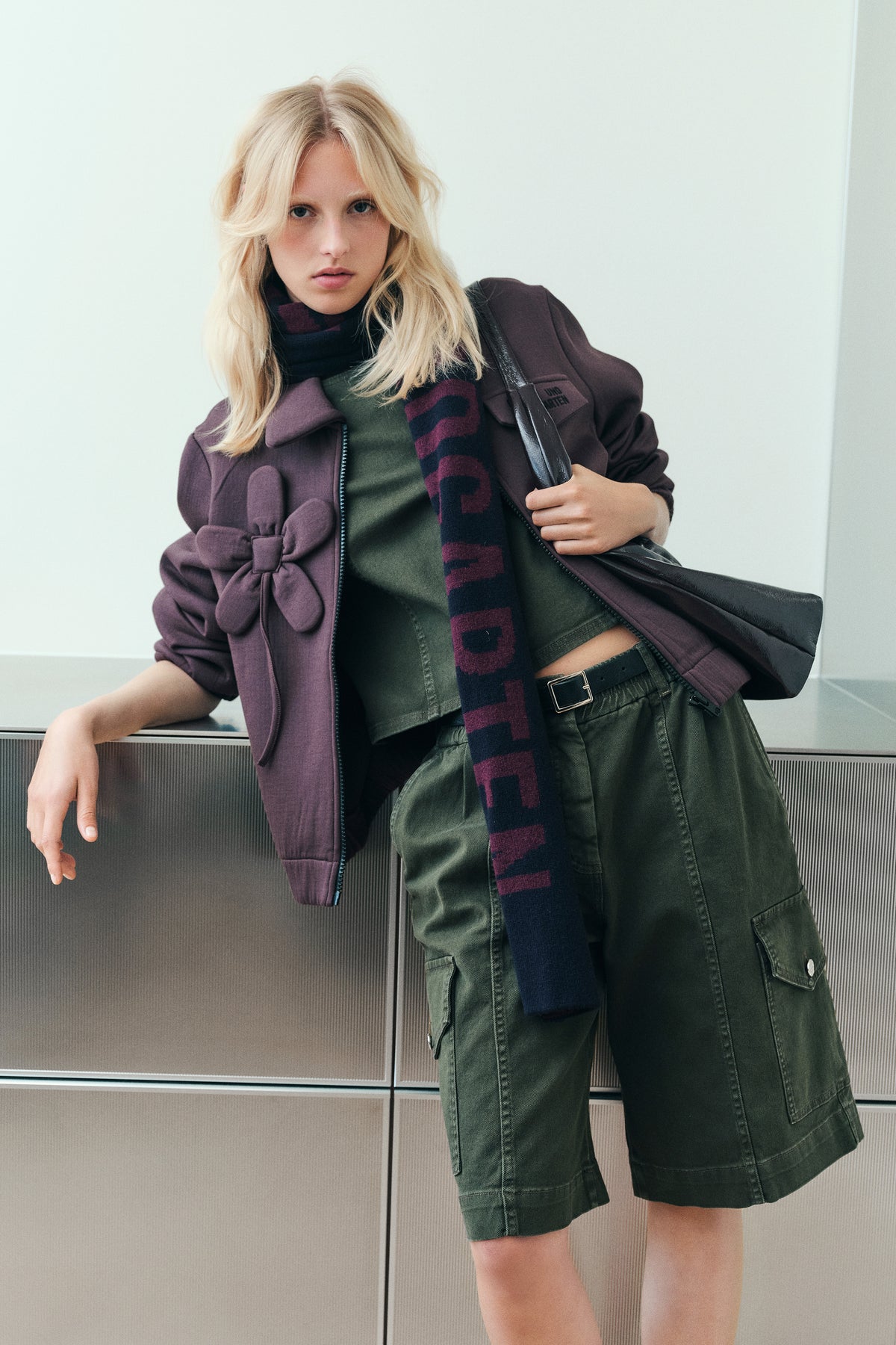 Campaign portrait featuring a plum belted jacket, olive top and cargo shorts, and a navy scarf with burgundy lettering.