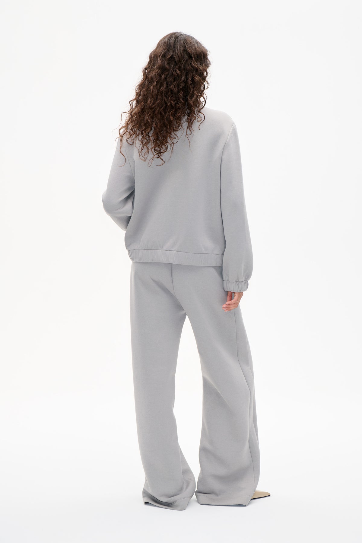 Achteraanzicht van een model in een lichtgrijs sweatshirt met rekbare manchetten en zoom, gecombineerd met een bijpassende broek met wijde pijpen.