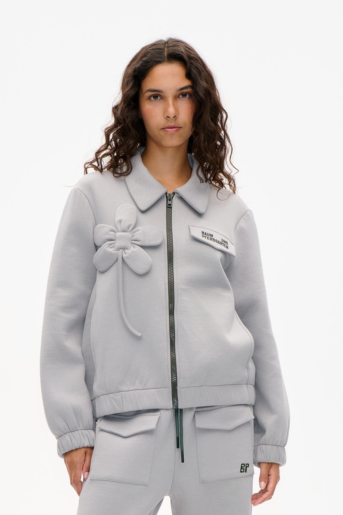 Model wearing a light gray zip-front jacket with an oversized collar, bow appliqué and chest patch.