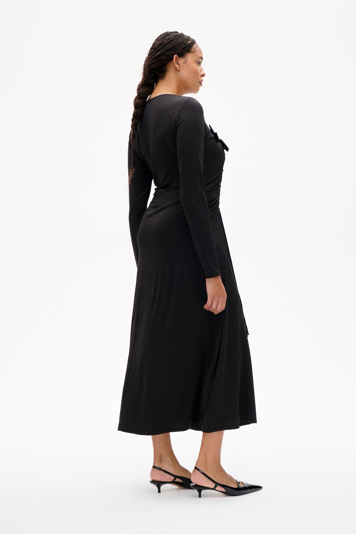 Side view of a model wearing a black long-sleeved dress with gathered side detailing and a bow accent.