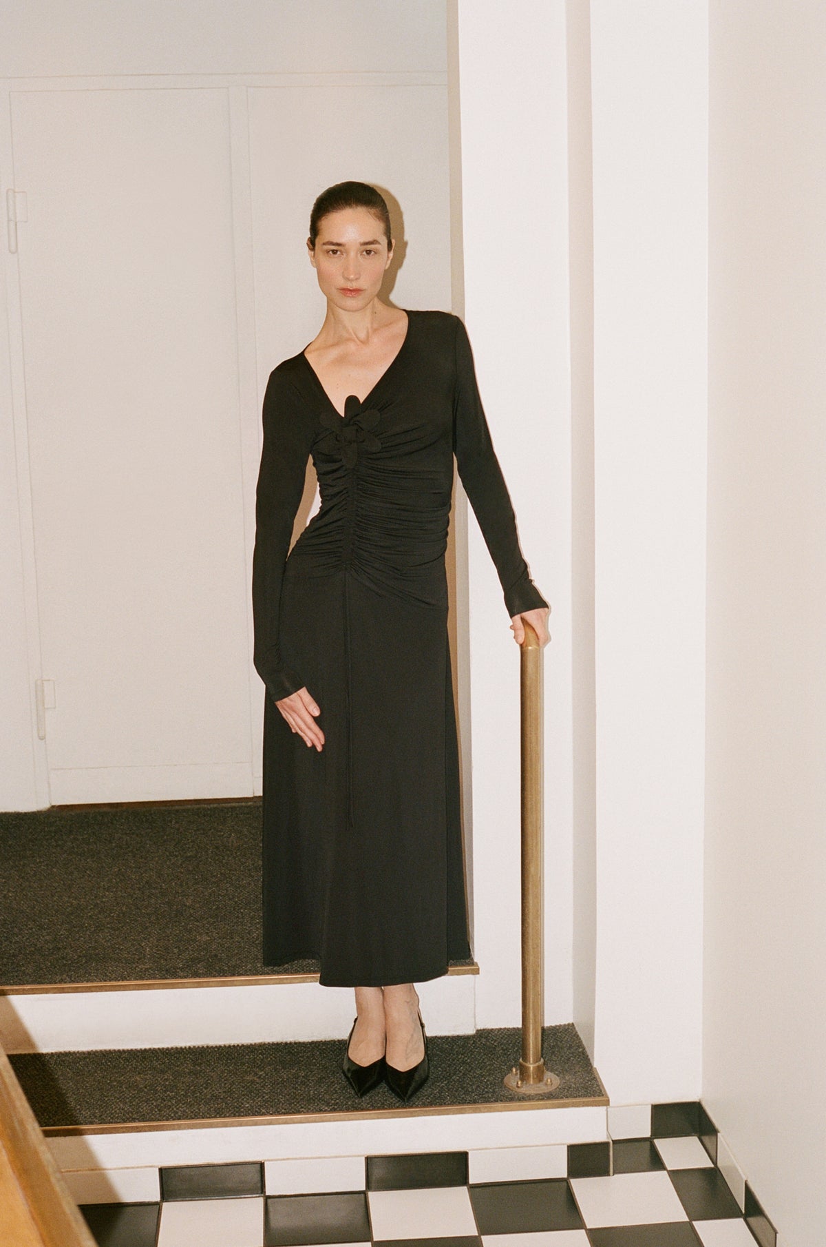 Model wearing a black long-sleeved maxi dress with a deep V-neck, decorative chest knot and gathered waist, standing on steps.