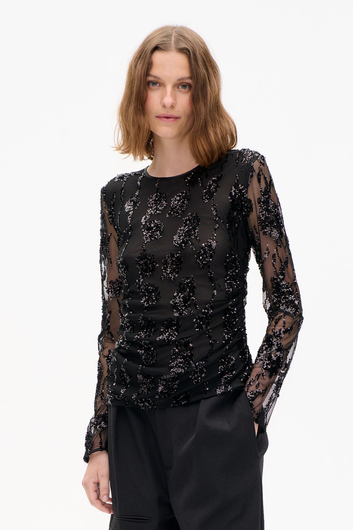 Front view of a model wearing a sheer black long-sleeved top with floral sequin embellishment and gathered waist detailing.