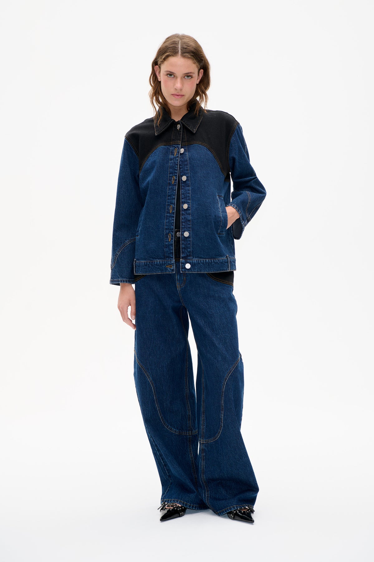 Model wearing a blue denim jacket with a contrasting black shoulder yoke, pointed collar and metal buttons, paired with wide-leg jeans.