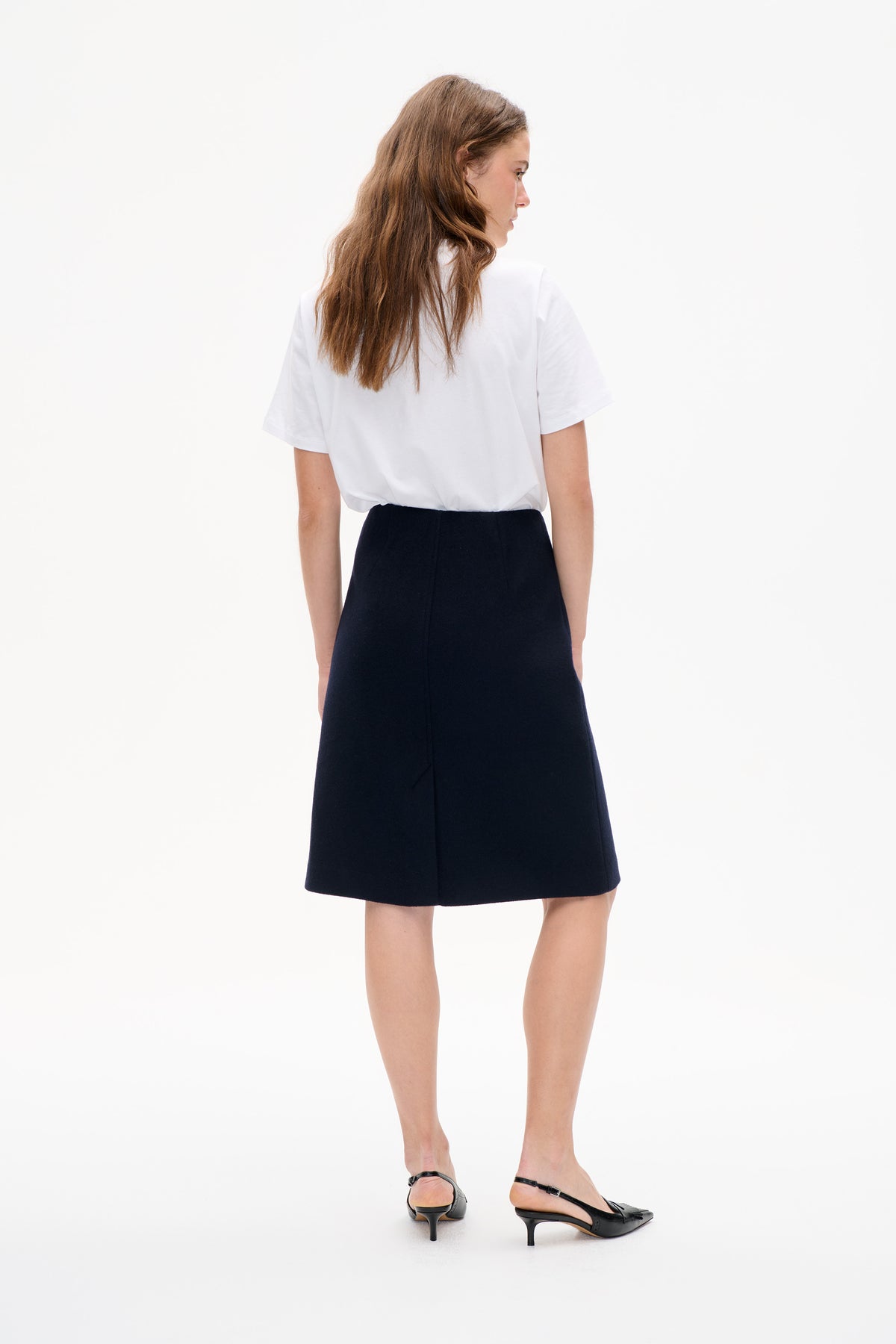 Model viewed from behind wearing a plain white short-sleeved T-shirt, black knee-length skirt and black slingback heels.