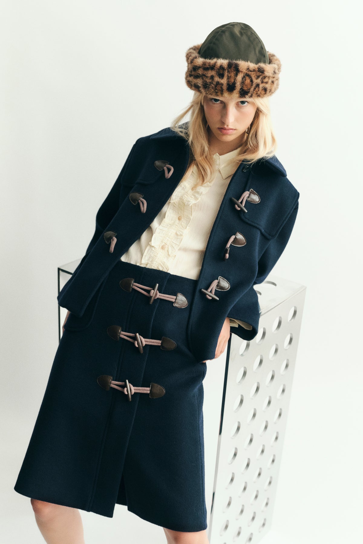 Campaign portrait of a model wearing a navy duffle coat with pink rope fastenings, a cream ruffled blouse and a brown patterned fur hat.