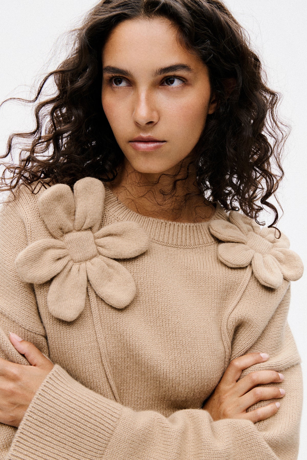 Close-up campaign portrait of a model wearing a beige rib-knit sweater with a round neckline and voluminous sleeves, arms folded.