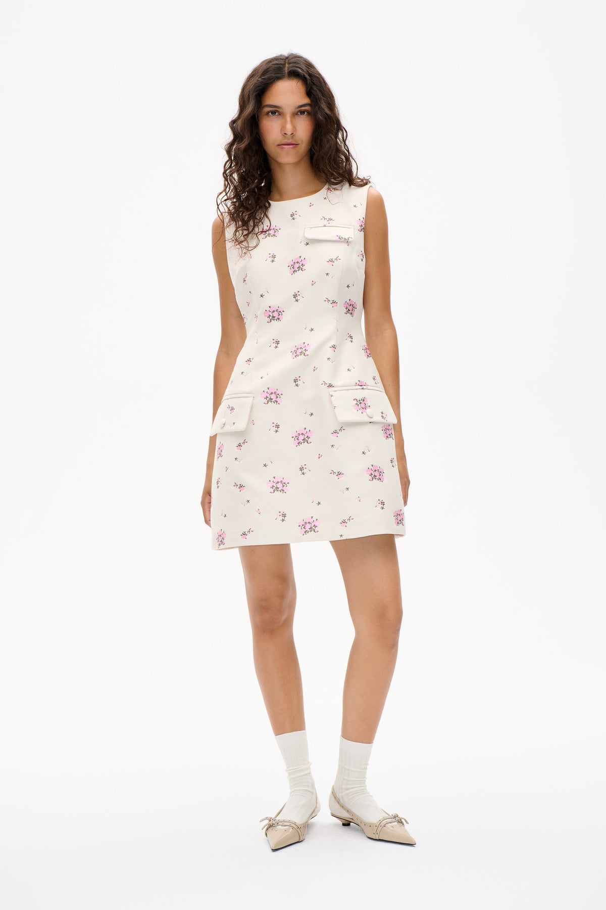 Model wearing a sleeveless ivory mini dress with scattered pink floral print and flap pockets, shown from the front.