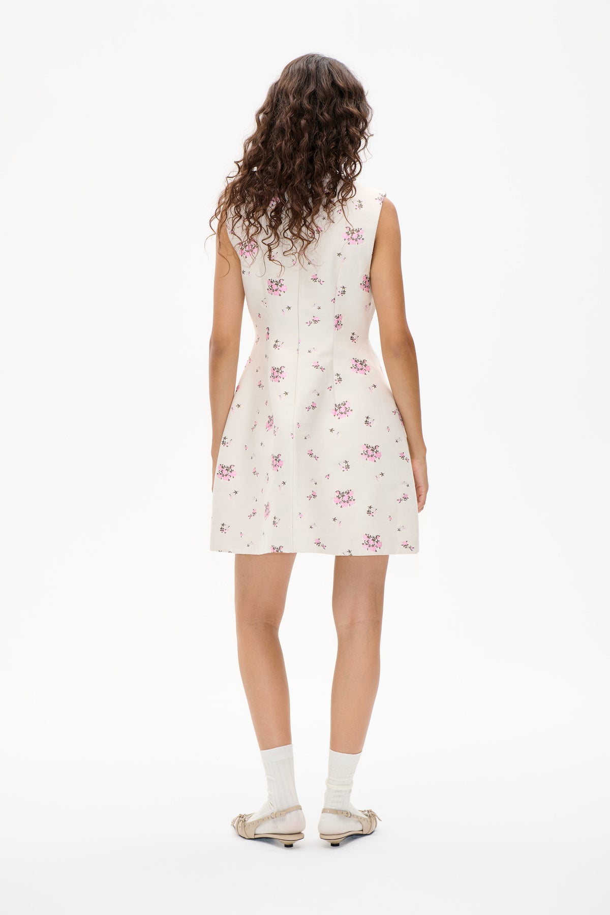 Back view of a cream sleeveless mini dress with scattered pink and brown floral print, styled with white socks and beige flats.