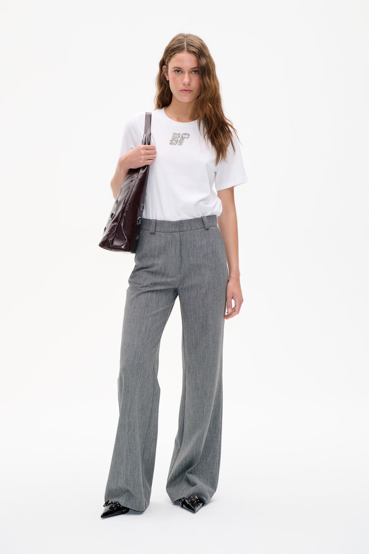 Model wearing a white short-sleeve T-shirt with jeweled initials and grey wide-leg trousers, carrying a glossy dark shoulder bag.