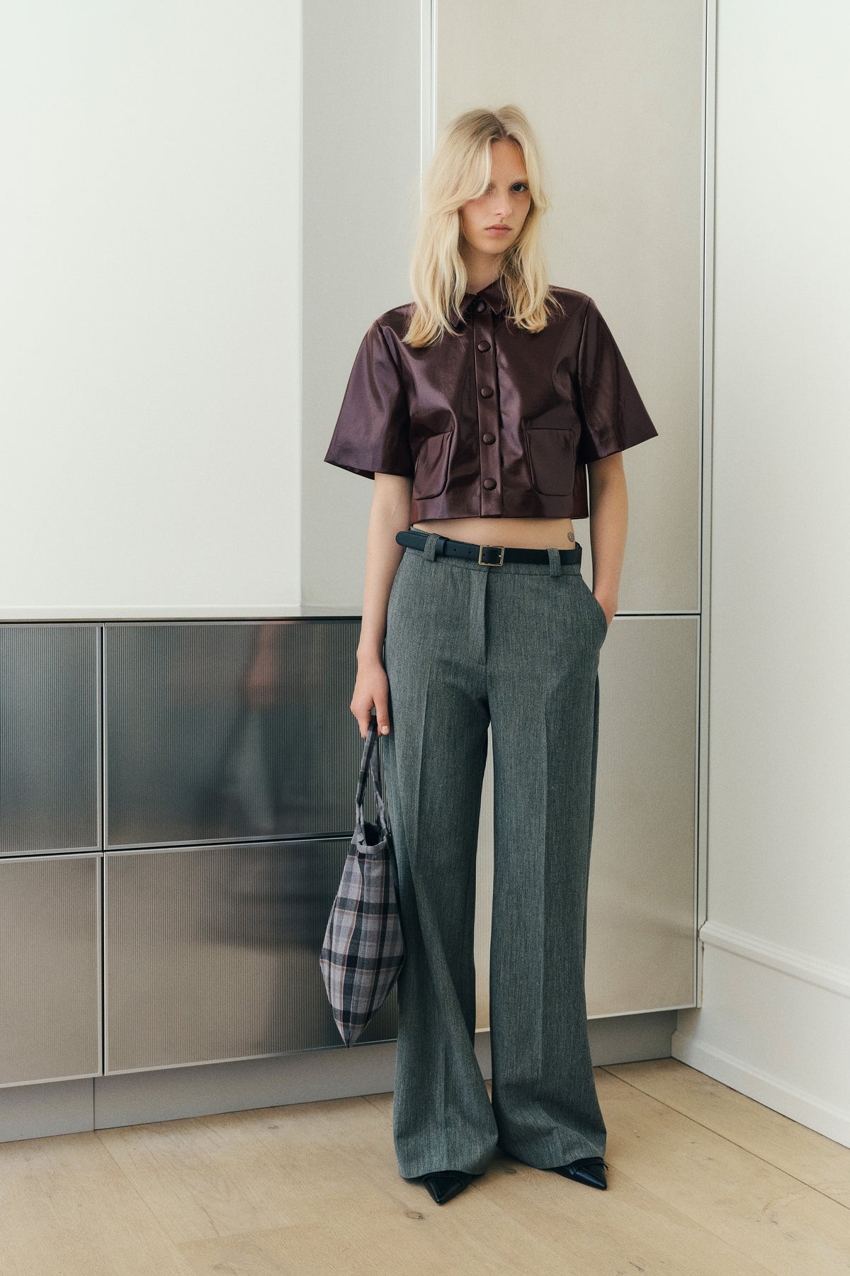 Portrait de campagne d'un mannequin portant une chemise courte boutonnée en satin brillant prune, un pantalon gris à jambes larges et un cabas à carreaux.