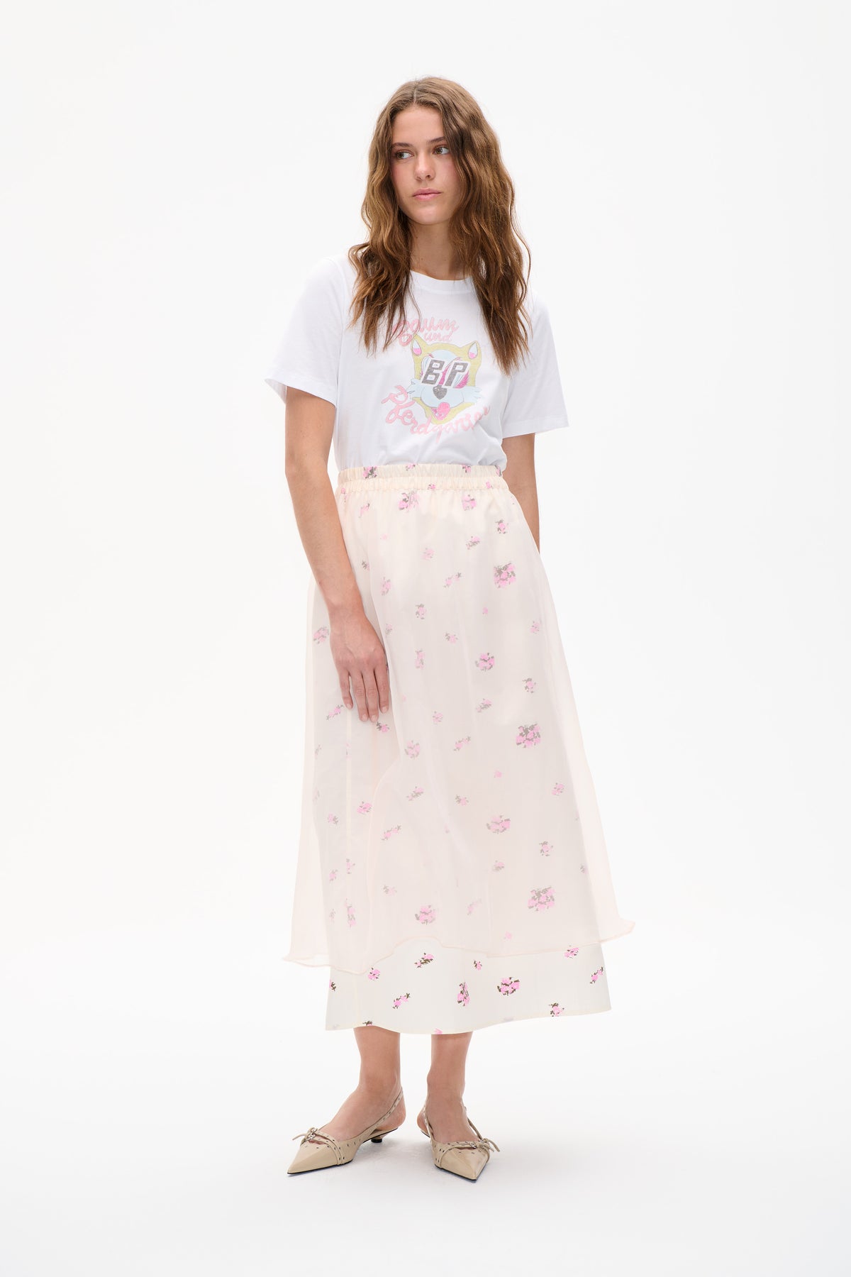 Model wearing a white graphic T-shirt with a colourful cat emblem and a sheer ivory floral skirt against a white studio backdrop.