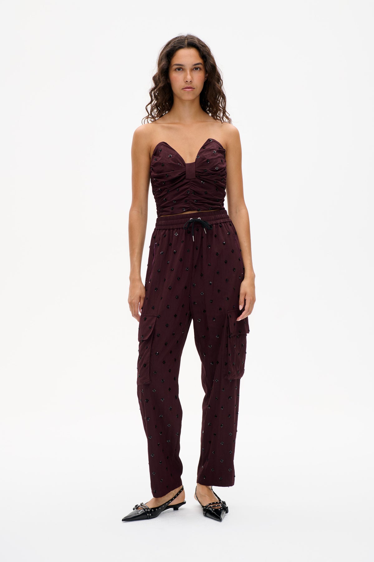 Front view of a model in a burgundy strapless ruched top and matching drawstring cargo trousers, both patterned with small dark floral embellishments.