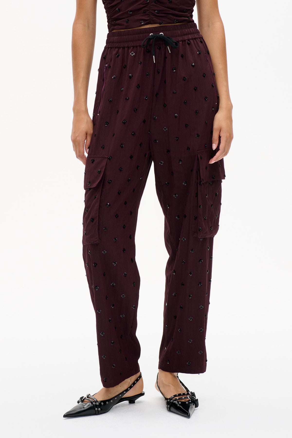 Front detail of burgundy drawstring trousers with scattered black floral embellishments, cargo pockets and tapered legs.