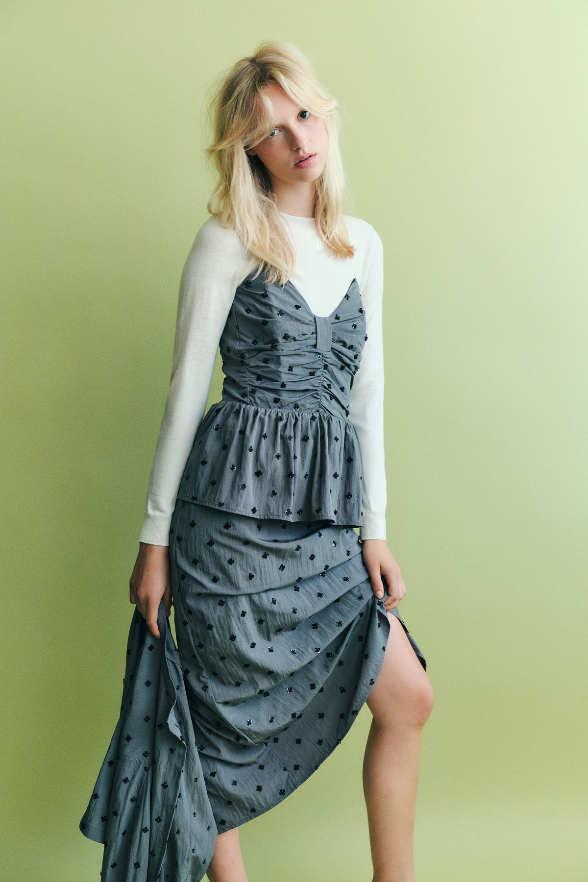 Model wearing a grey dress with a ruched bodice and black floral embellishments over a white long-sleeved top against a pale green background.