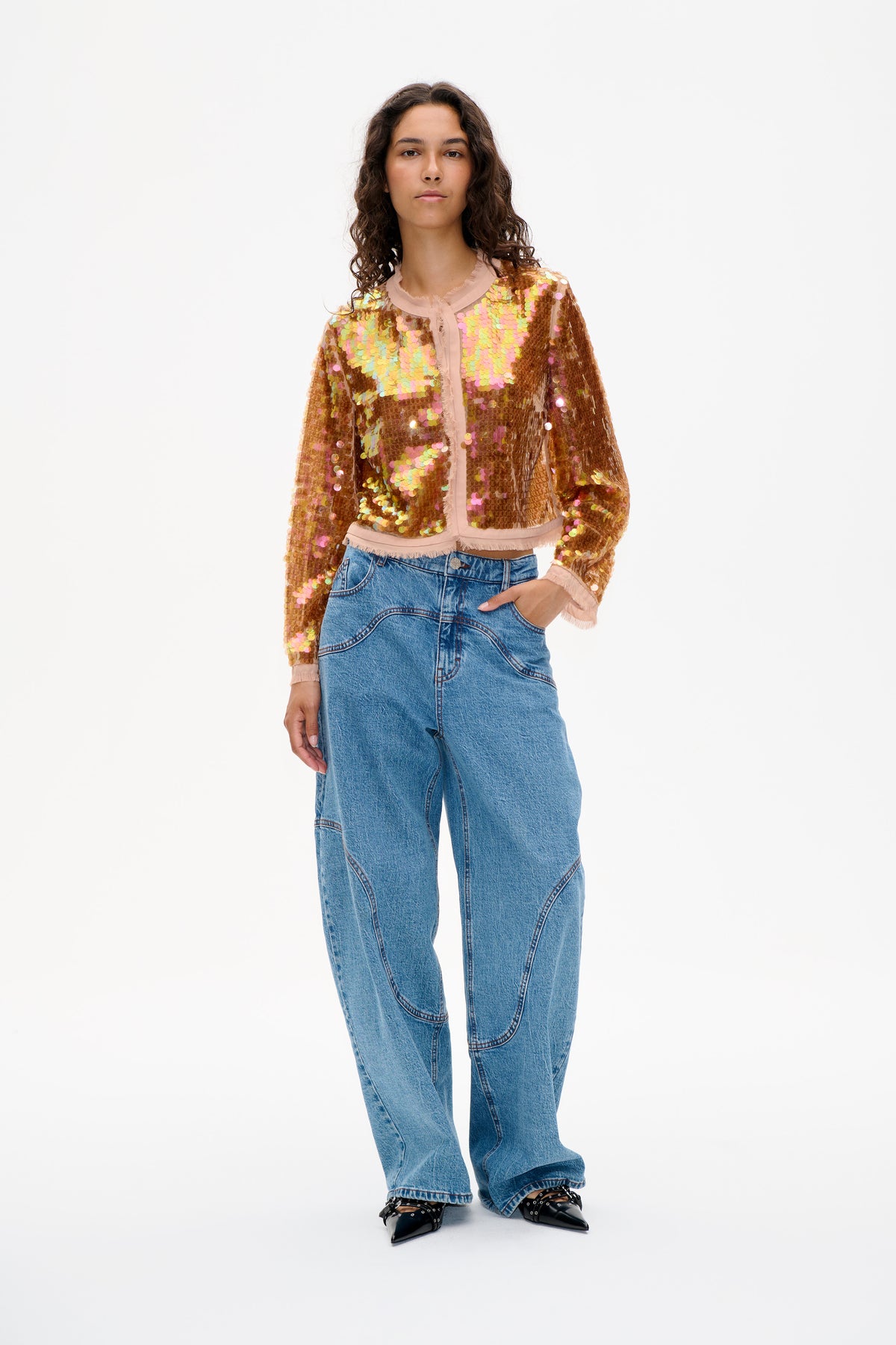 Model wearing a bronze-gold iridescent sequin jacket with pale pink trim and wide-leg blue jeans, shown from the front.