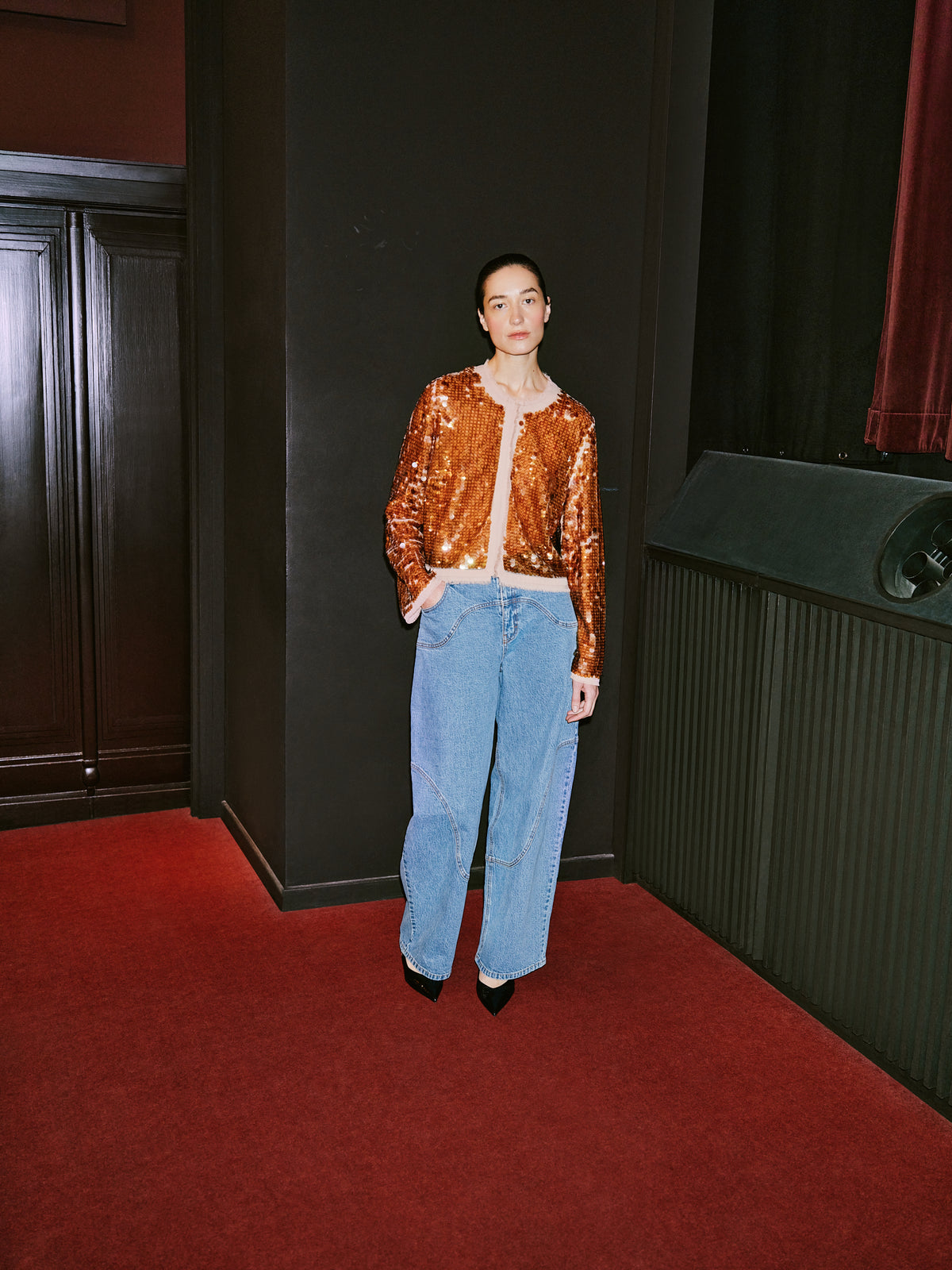 Model wearing a copper-orange sequined cardigan with pale trim and light-wash wide-leg jeans in a dark interior.