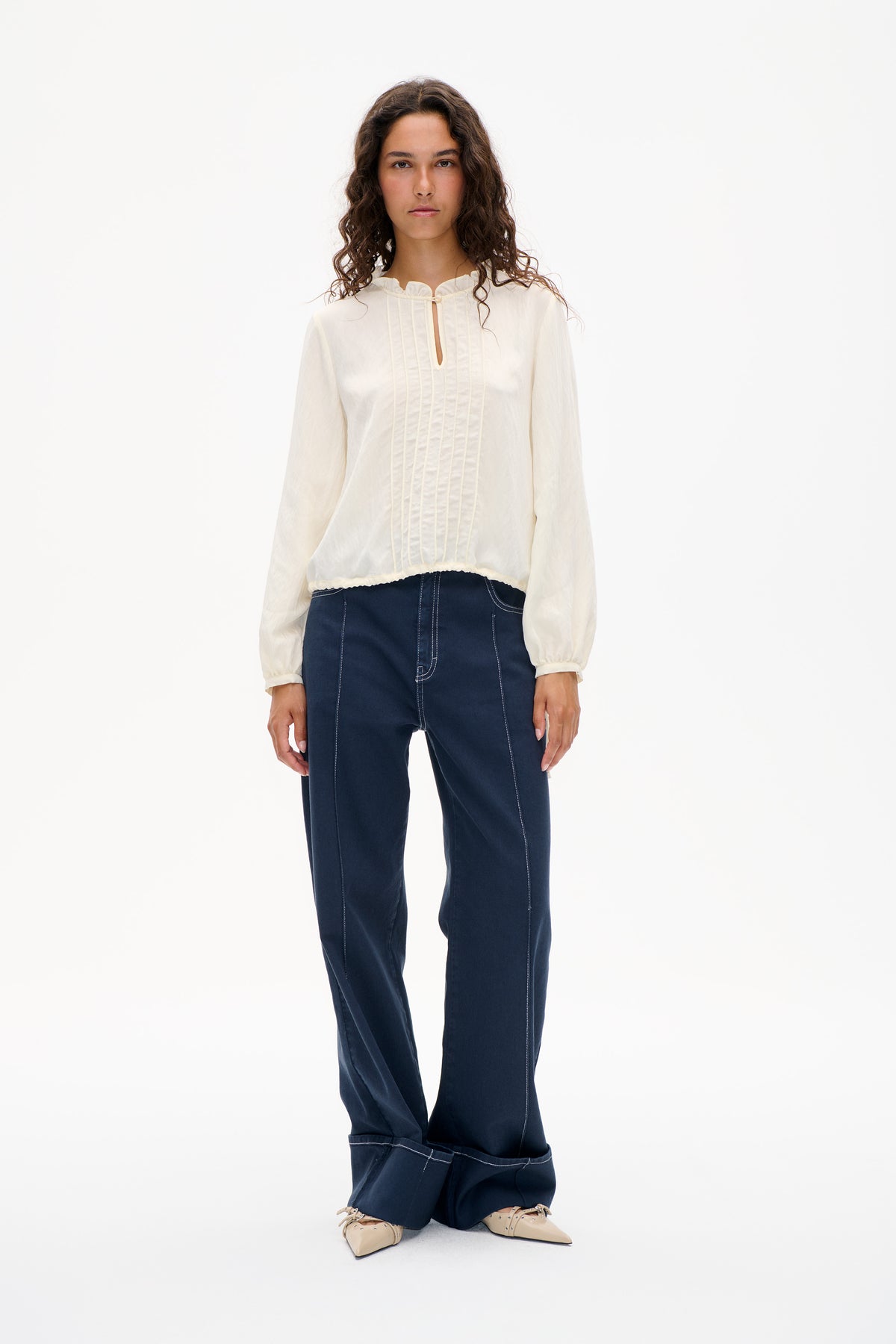 Vorderansicht eines Models, das eine elfenbeinfarbene, langärmlige Bluse mit gerüschtem Ausschnitt, Biesen und kleinem Schlüssellochdetail trägt, kombiniert mit dunkelblauen Jeans.