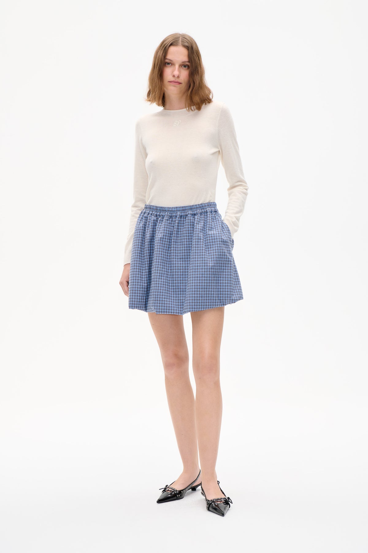 Model wearing an ivory crewneck sweater with a small chest monogram, a gathered blue checked mini skirt and black pointed flats against a white background.