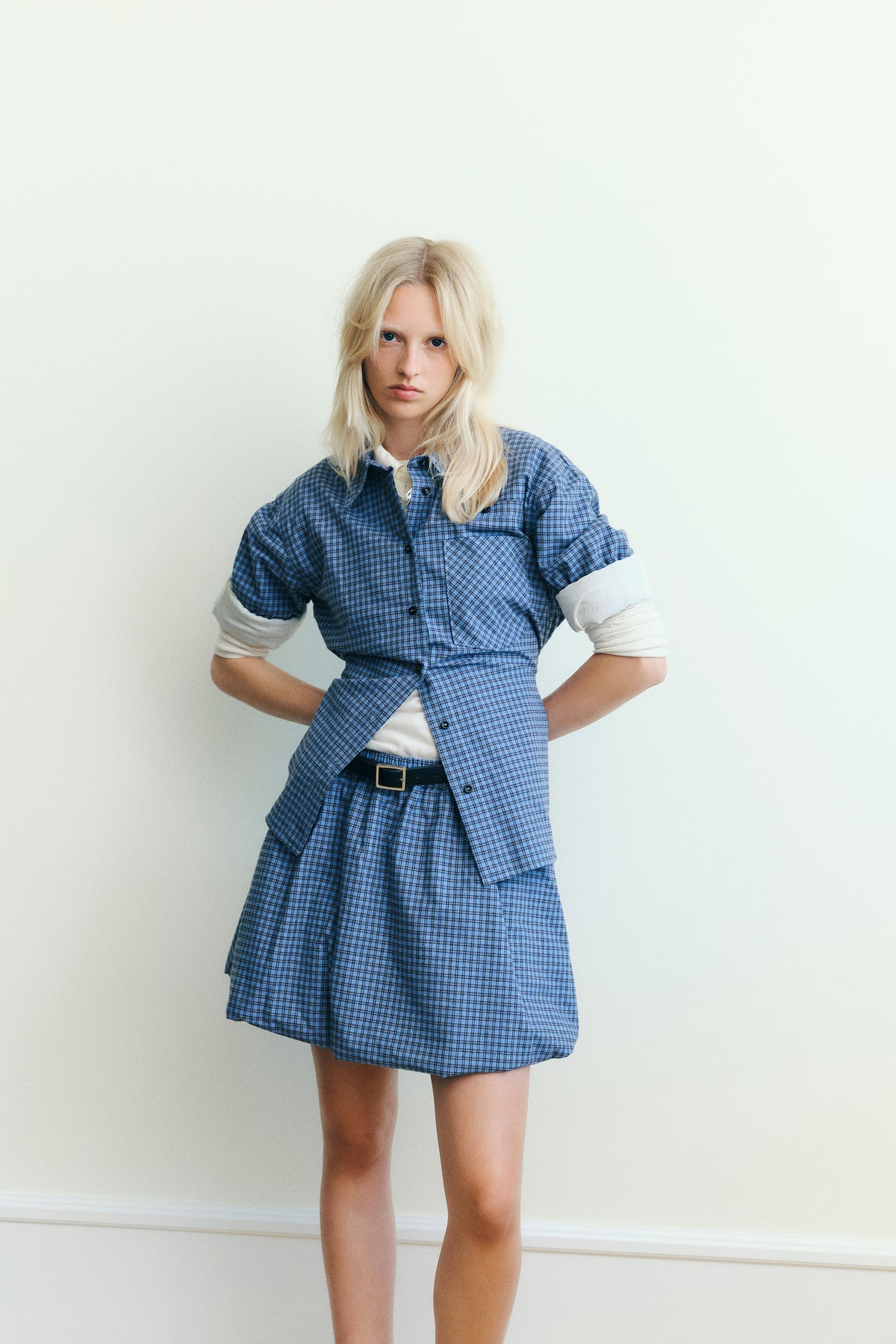 Campaign portrait of a model wearing a blue checked button-front shirt over a cream top with a matching skirt against a pale backdrop.