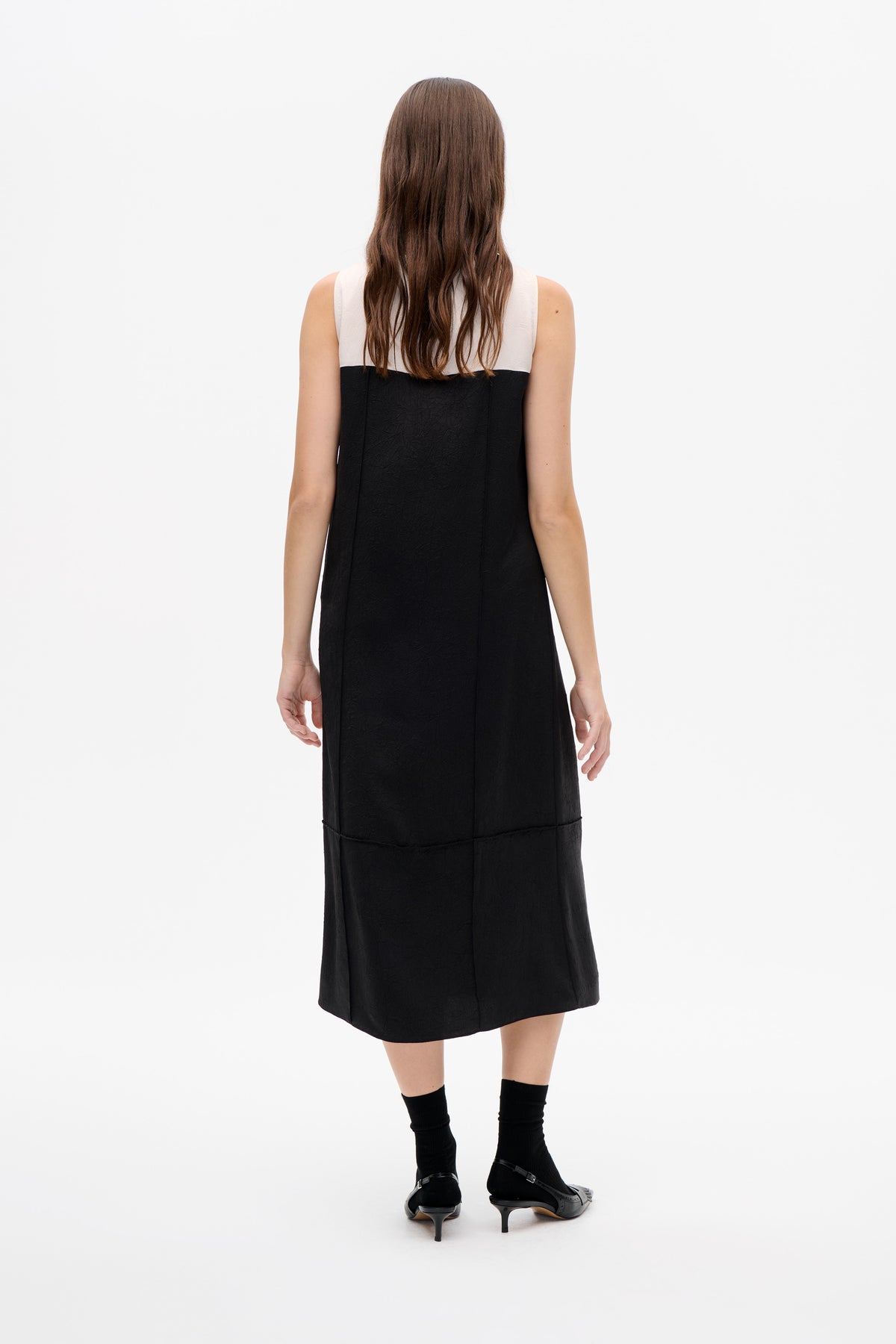 Back view of a sleeveless black midi dress with a white upper panel and subtle textured fabric, modeled with black socks and heels.