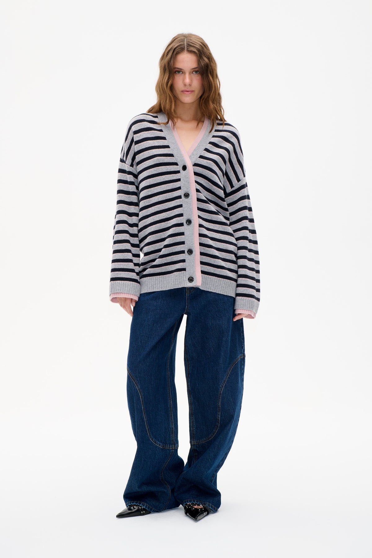 Model trägt einen grauen Cardigan mit V-Ausschnitt, Knopfleiste und Streifen in marineblau und hellrosa, kombiniert mit dunkelblauen Jeans.