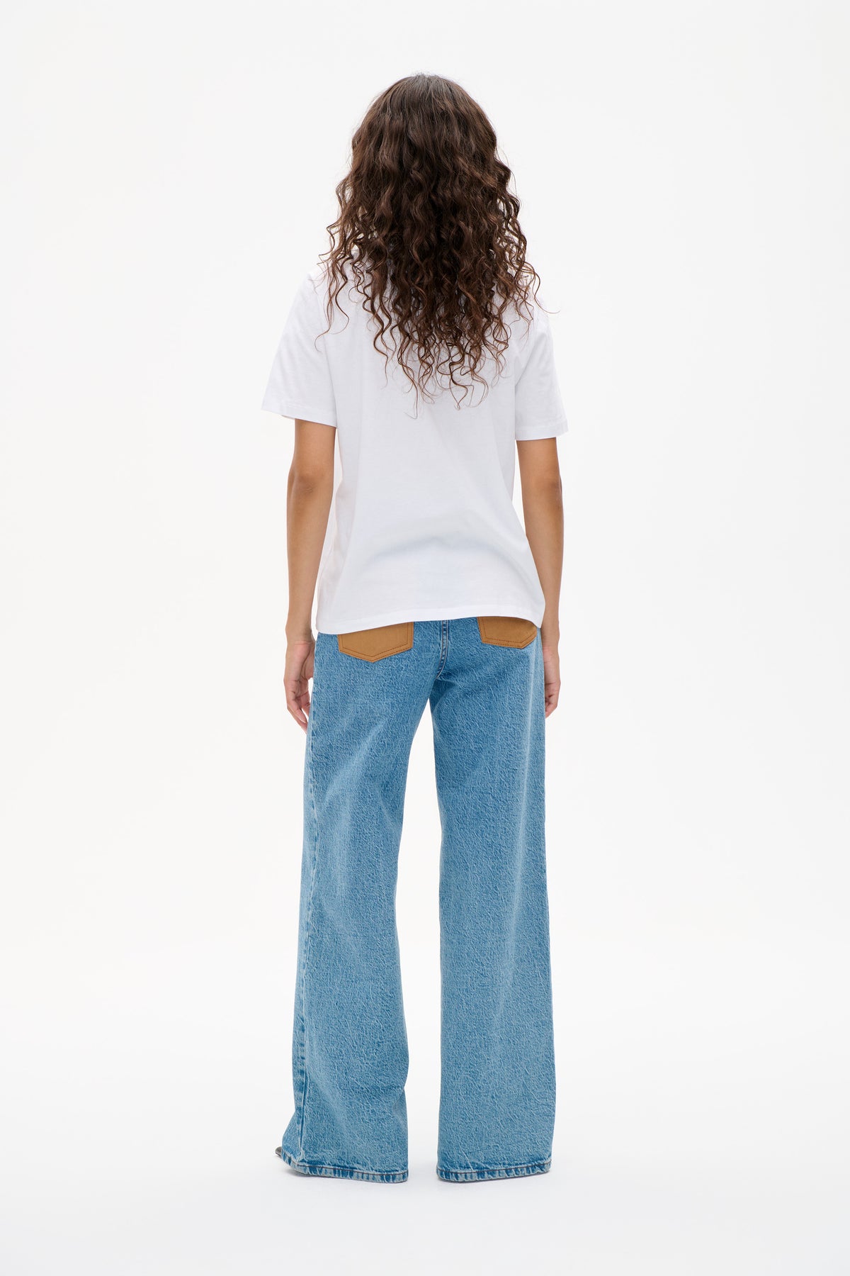 Achteraanzicht van een model in een effen wit T-shirt met korte mouwen, lichtblauwe wijde jeans en lichtbruine achterzakken.