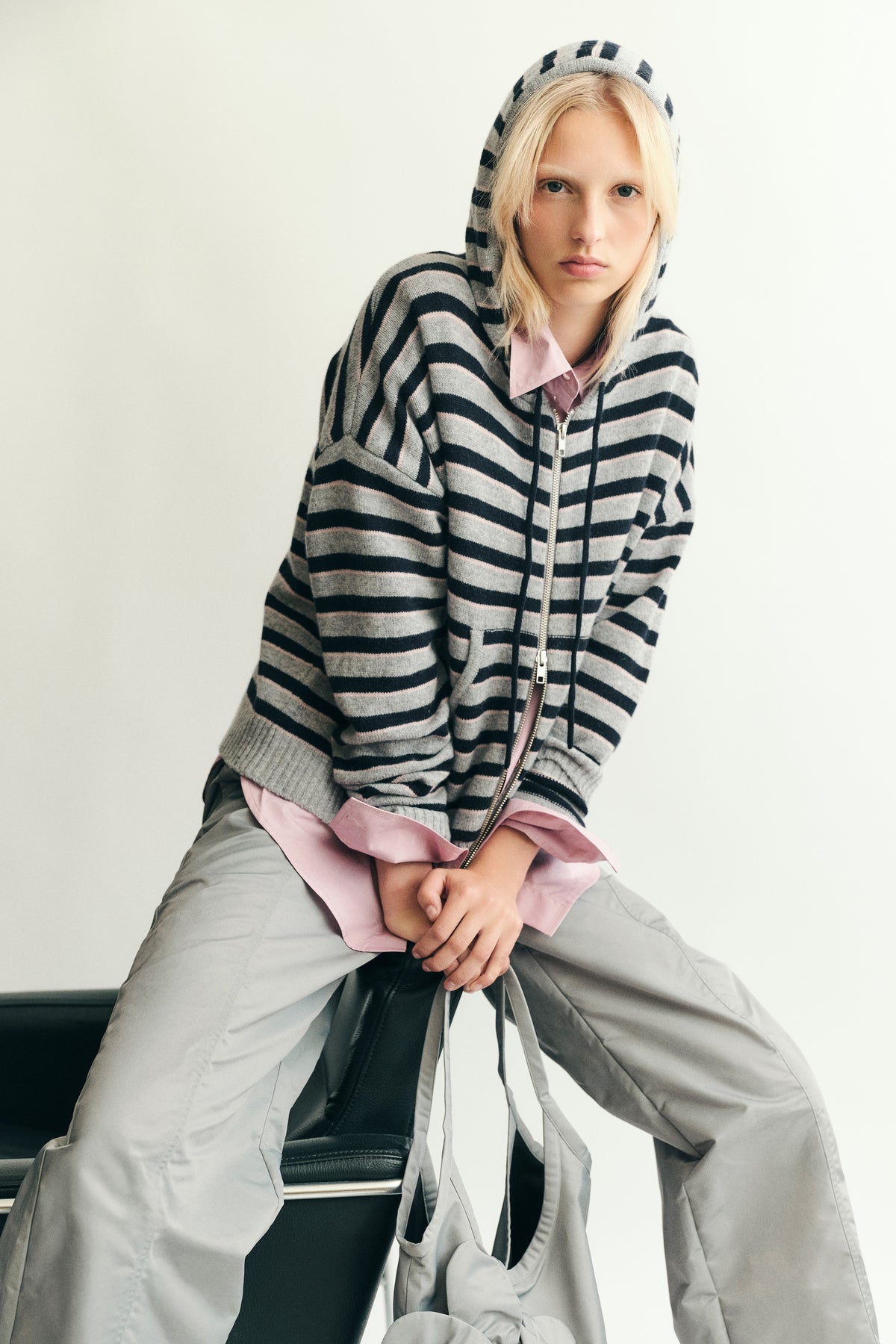 Seated model wearing a grey hooded zip-up sweater with navy and pale pink stripes over a pink shirt and wide grey trousers