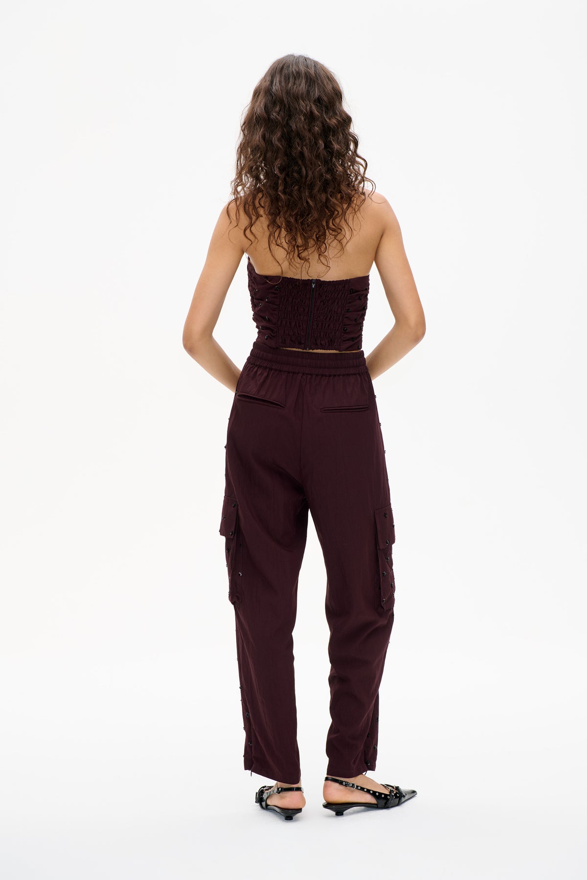 Back view of a model wearing a burgundy strapless smocked top with a center zipper and matching wide-leg trousers with black embellishments.