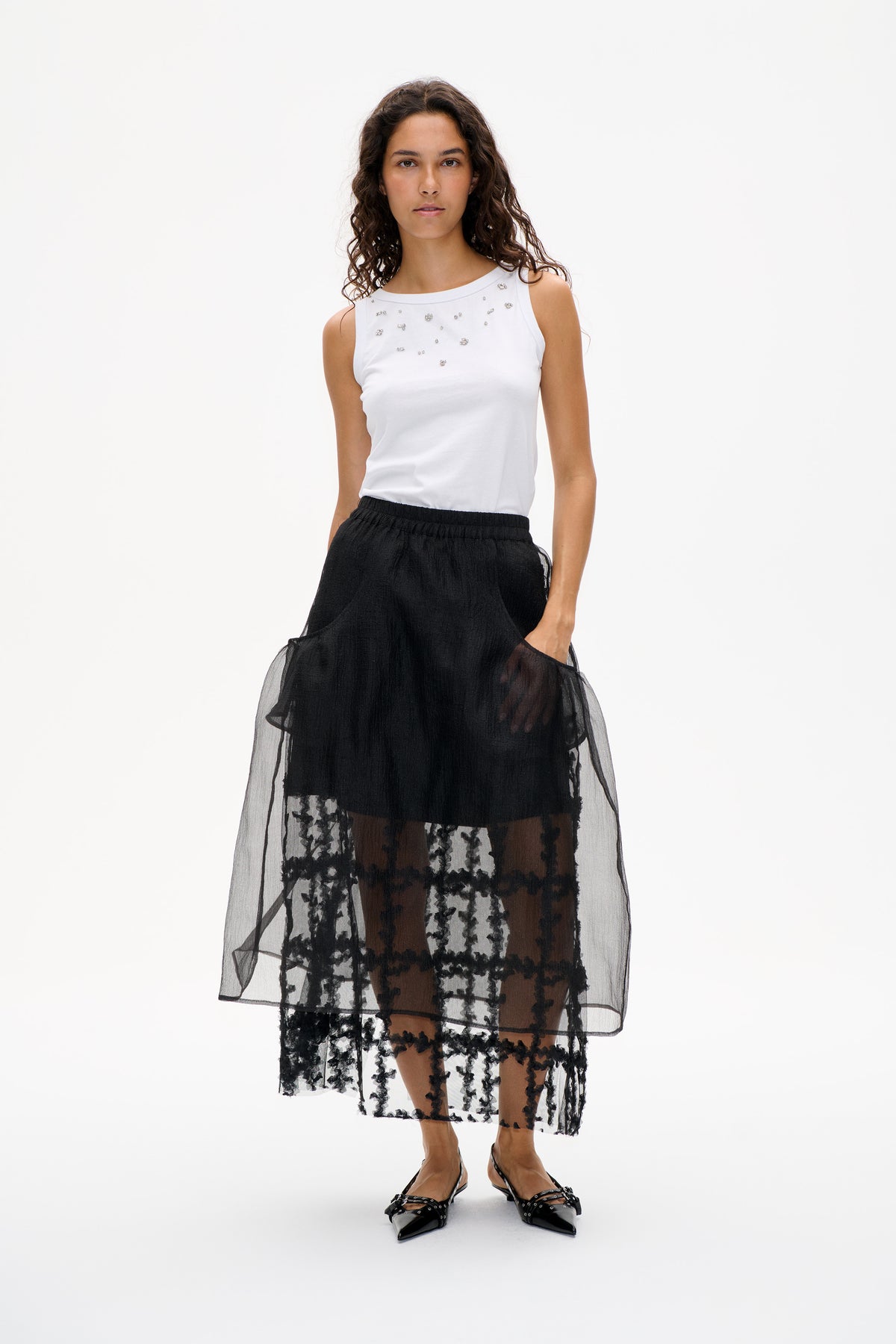 Model wearing a white sleeveless top with silver embellishments and a sheer black floral-patterned midi skirt.