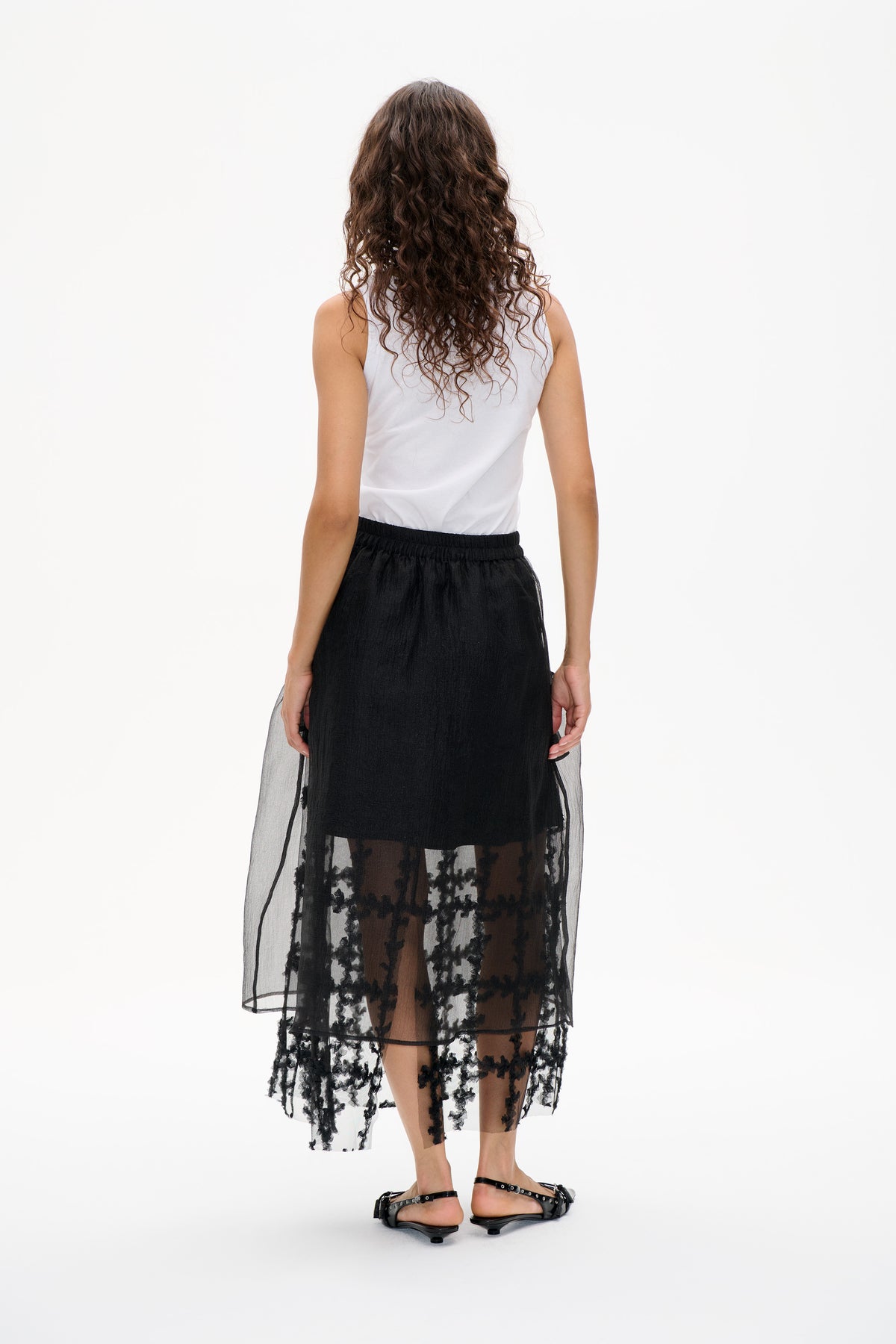 Rear view of a model wearing a white sleeveless top and a black skirt with a sheer floral-patterned overlay.