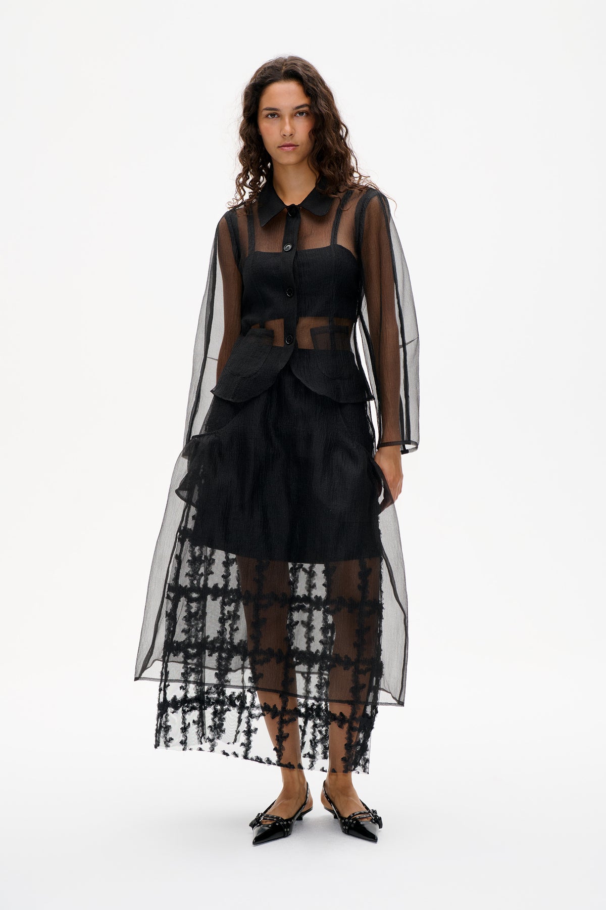 Model in a sheer black button-front shirt dress with a wide collar, layered over a black top and black-and-white patterned lace skirt.