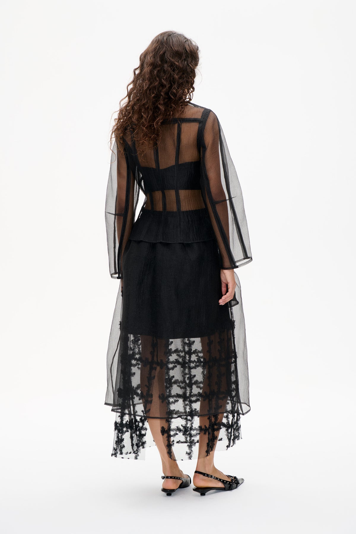 Back view of a model in a sheer black long-sleeved dress with geometric trim and floral detailing near the hem