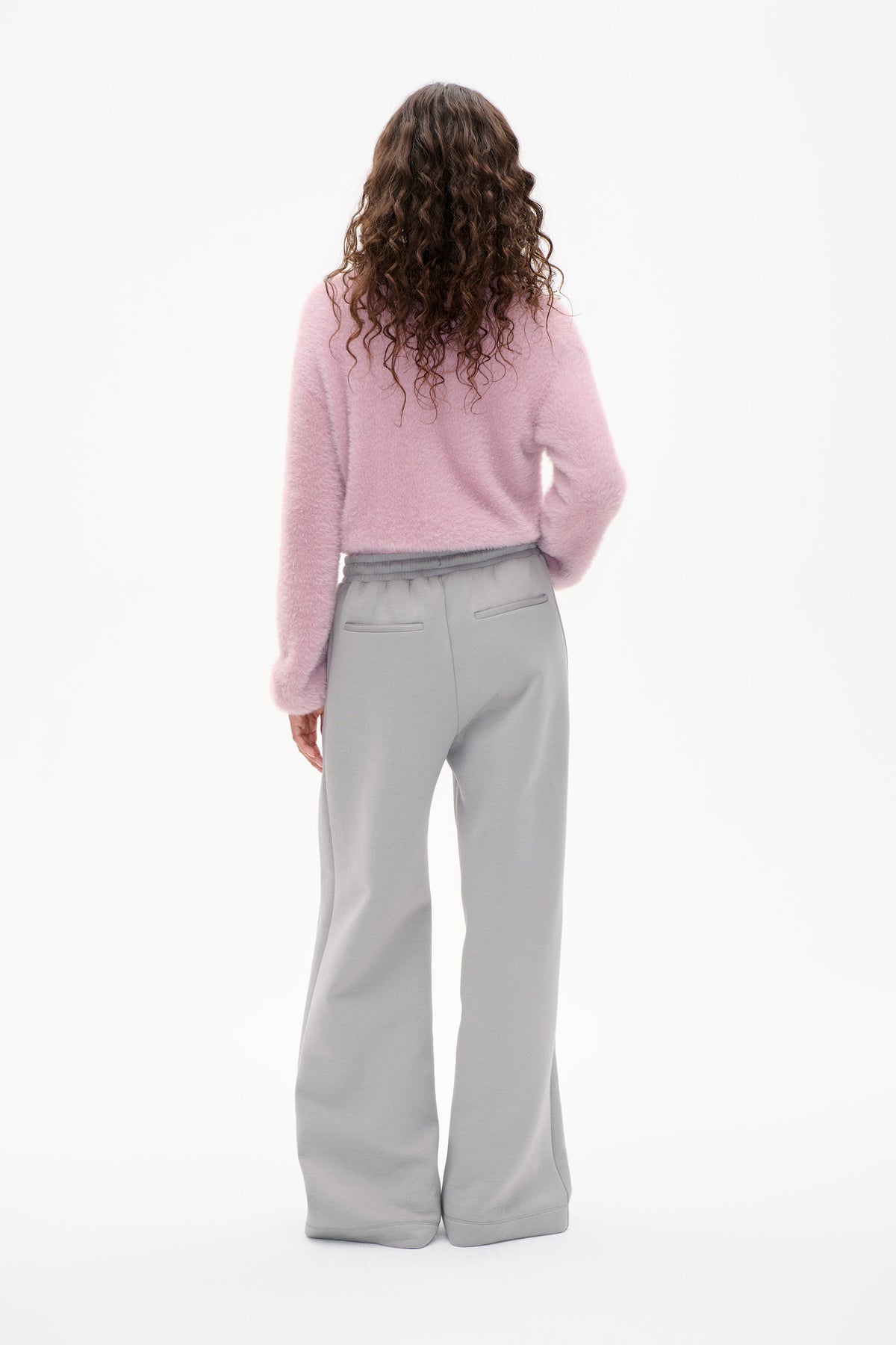 Back view of a model wearing a fuzzy pale pink sweater and light grey wide-leg trousers with an elasticated waistband and welt pockets.