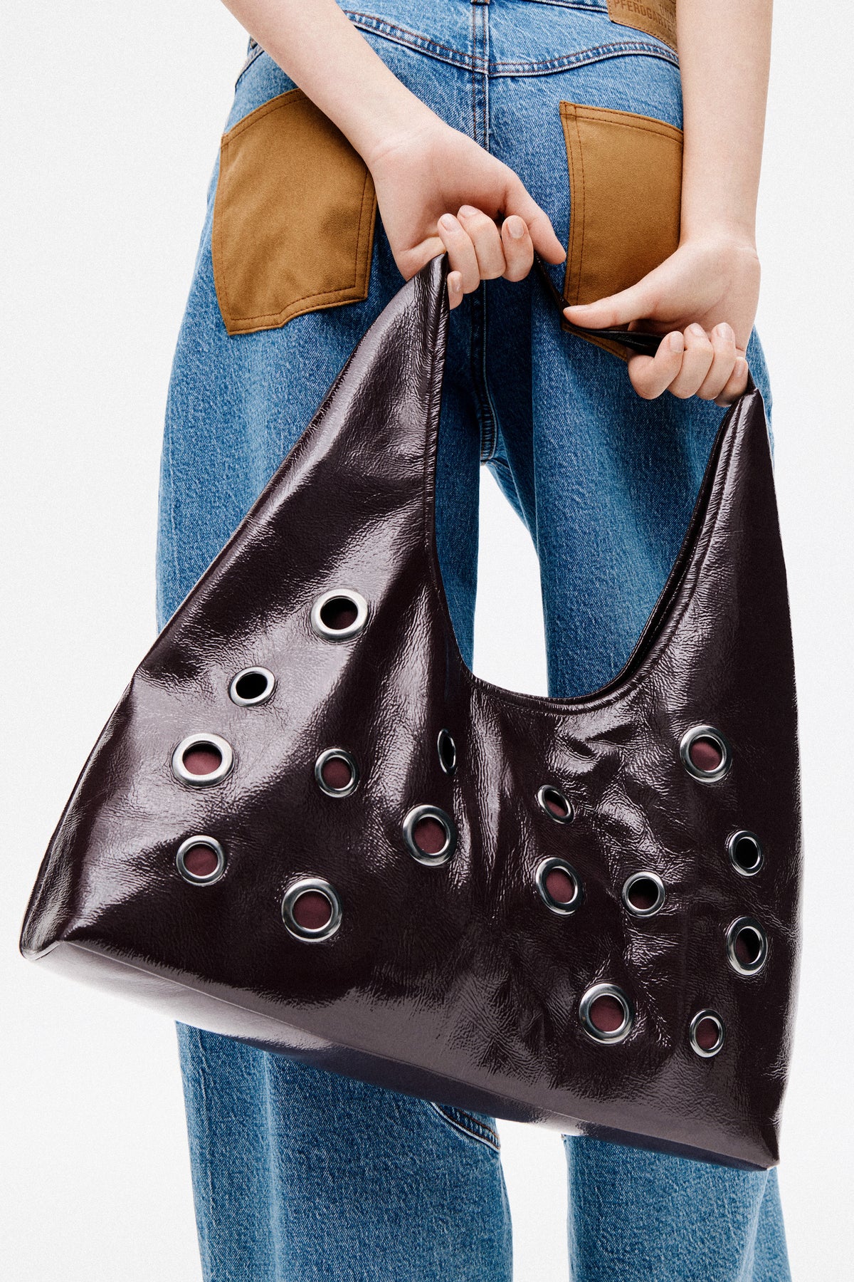 Close-up campaign view of blue jeans with tan patch pockets beside a glossy dark brown tote with metal eyelets.