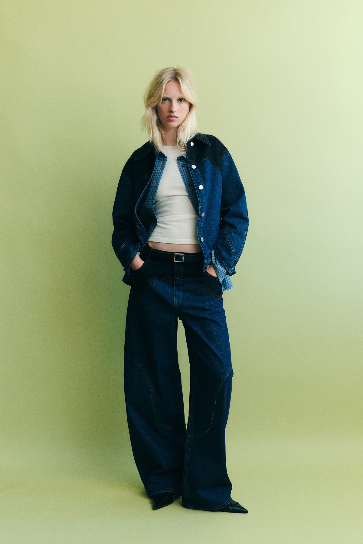 Model draagt een denimjas in donker marine en blauw over een blauw geruite blouse en een wit topje, gecombineerd met jeans met wijde pijpen tegen een lichtgroene achtergrond.