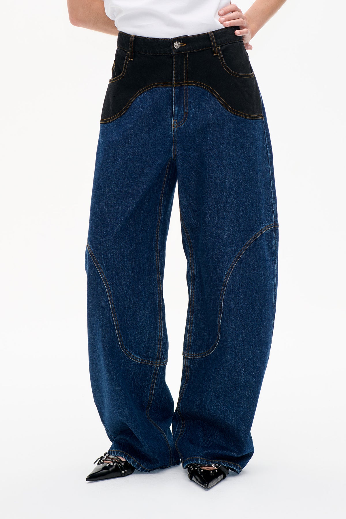 Detail van de voorkant van wijde blauwe jeans met zwarte gebogen heuppanelen, goudkleurige contrasterende stiksels en knoopsluiting.