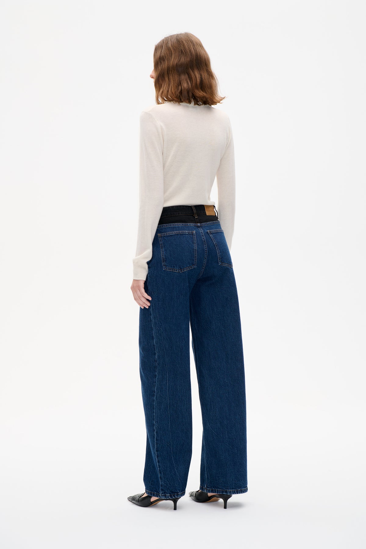 Back view of a model wearing dark blue wide-leg jeans with contrast stitching and a cream long-sleeve top.